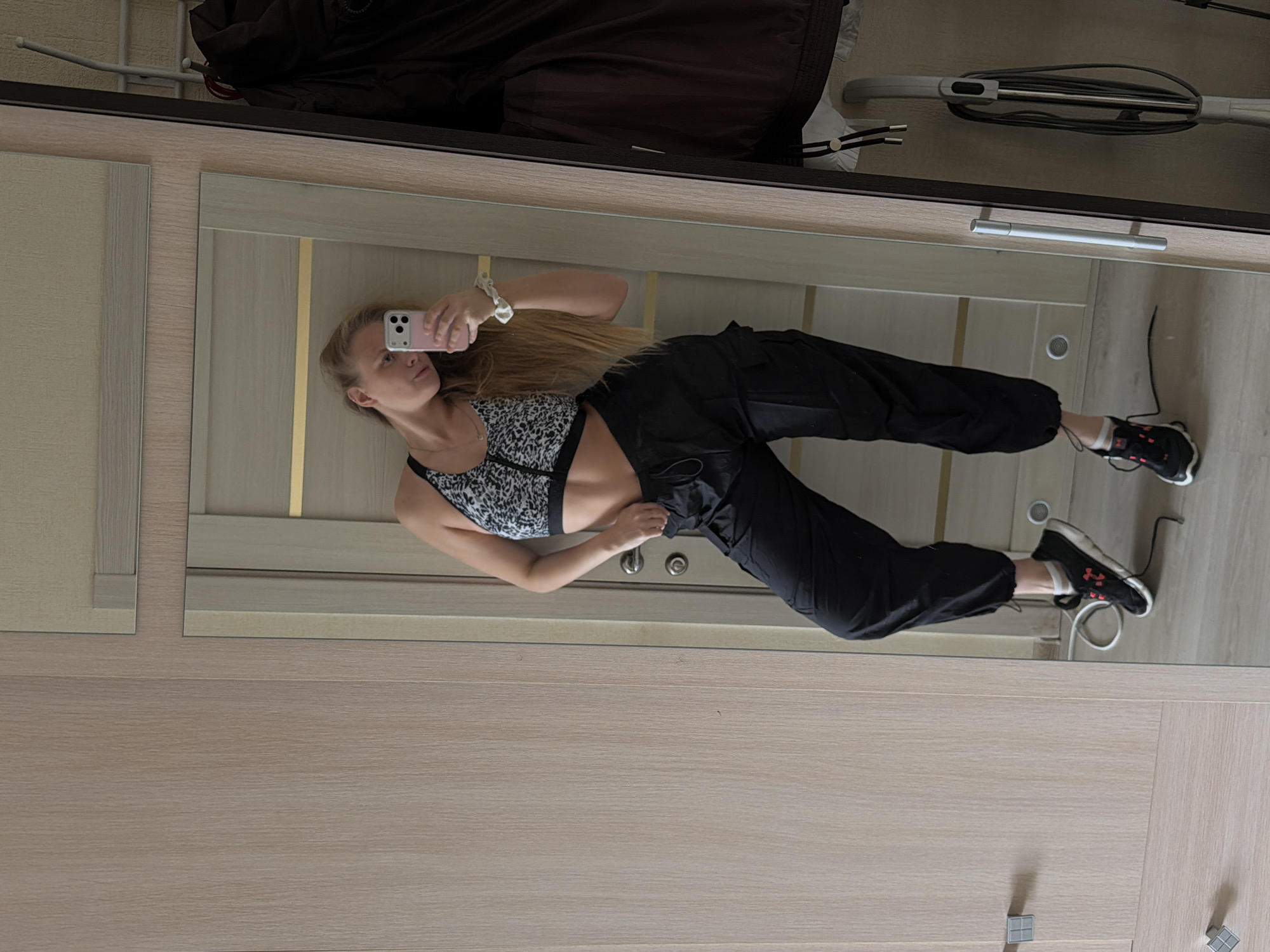 A young woman stands in front of a mirror, using a smartphone to take a selfie. She is wearing a leopard-print sports bra and black cargo pants. Her long blonde hair falls over her shoulder. She is standing in what appears to be a bedroom or dressing area, with a wooden cabinet behind her. A vacuum cleaner is partially visible at the top of the frame. The location is Izhevsk, Russia.