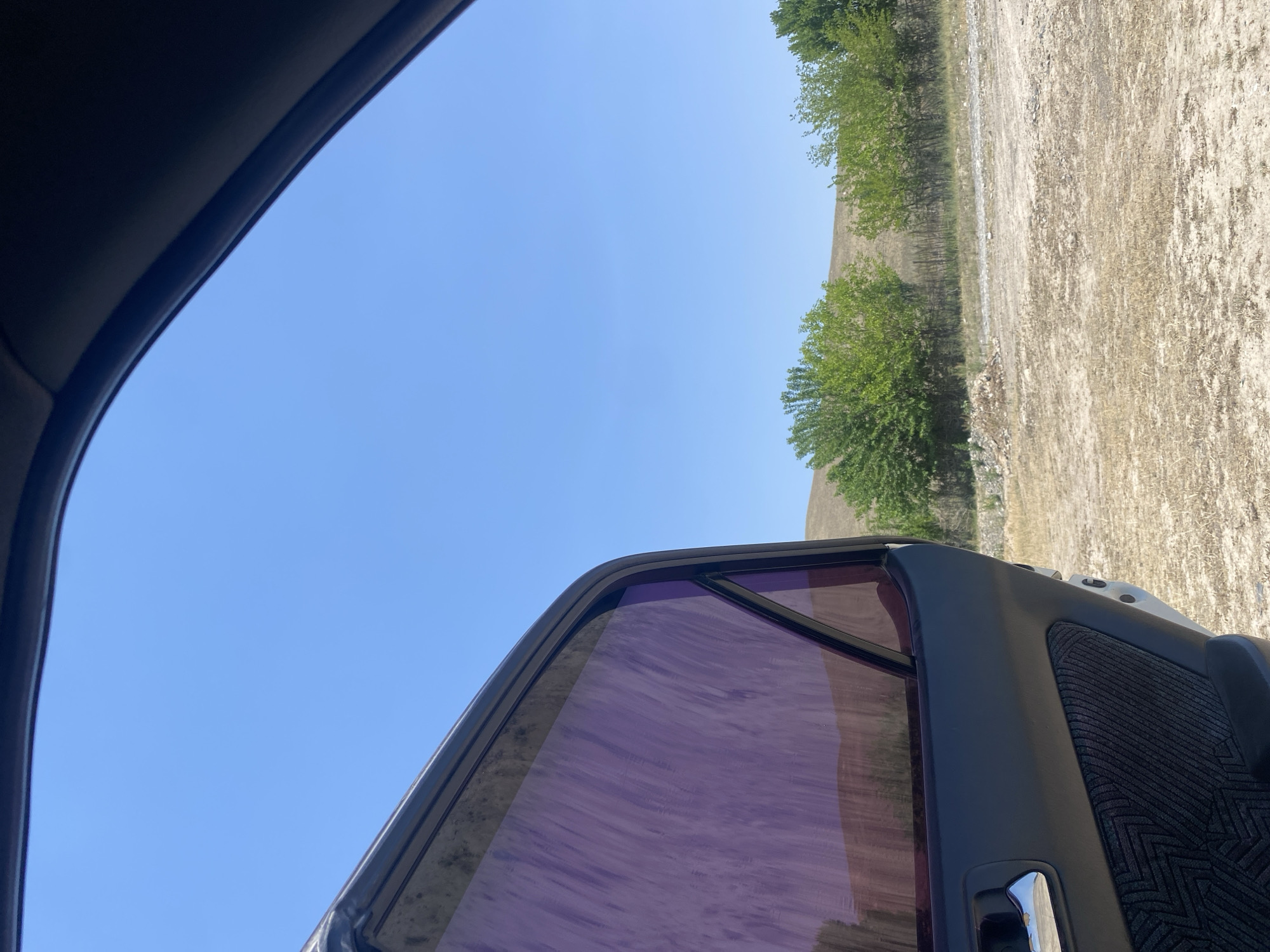 The image shows an open car door with purple-tinted windows, looking out onto a dry, dusty landscape under a clear blue sky. In the background, there are sparse trees and rolling hills characteristic of the region. This scene is consistent with the natural environment found near Naray, Kyrgyzstan, where such arid terrain and vegetation are common. There are no people visible in the image.