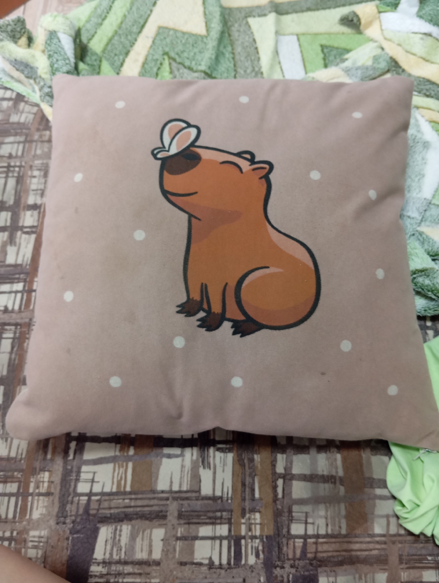 A close-up, medium shot shows a light brown pillow with a cartoonish brown capybara with a white butterfly on its head. The capybara is depicted with its eyes closed and a smile, sitting with its front paws together. The pillow is decorated with scattered white polka dots. The pillow rests on a surface covered by a patterned blanket or bedspread with green, yellow, and white abstract geometric shapes. To the left and right sides of the pillow, the background shows a brown and gray checkered pattern. A bright green fabric is visible bunched up on the right side of the image. The scene appears to be an indoor setting. There is no indication of the location being Novodesyatnikovo, Russia.