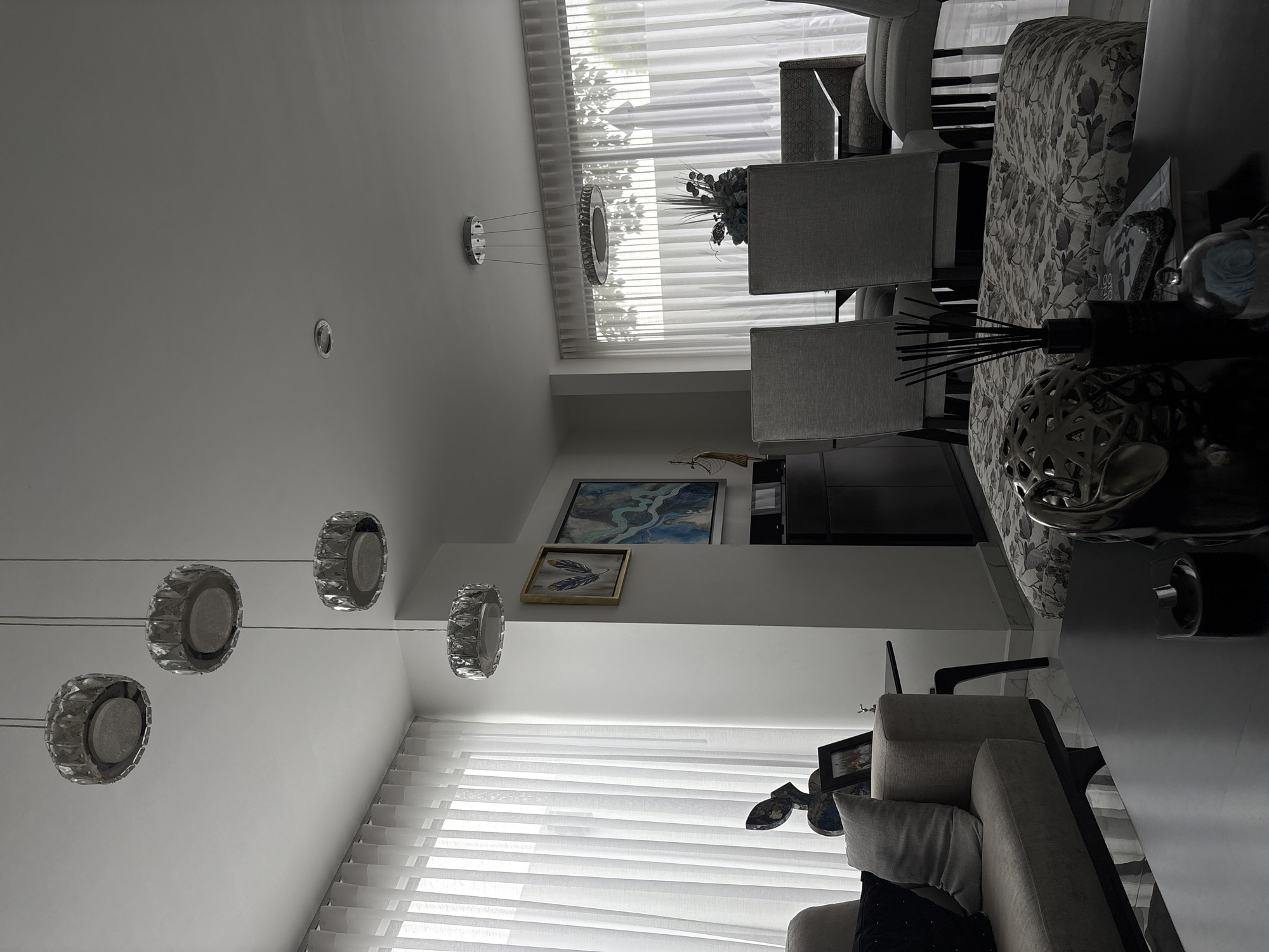 The image depicts a modern, well-lit living and dining area, likely located in San Bernardino Tlaxcalancingo, Mexico. The room is furnished with contemporary pieces, including a sectional sofa with throw pillows, a dark coffee table with decorative items like an ornamental sphere, a glass dome, and a reed diffuser, and a dining set with upholstered chairs and a long table. A patterned ottoman is positioned in the foreground. The space is characterized by large, floor-to-ceiling white curtains that diffuse the natural light entering the room. Several pendant lights hang from the ceiling; a series of four circular, crystal-accented lights are prominent over the living area, and a larger, ring-shaped light fixture is suspended over the dining table. Artwork adorns the wall, including a framed piece with blue feathers and a larger abstract painting. A dark cabinet is visible against the wall. The flooring appears to be a polished marble. There are no people present in the scene. The overall atmosphere is one of sophisticated, minimalist design and comfortable living.