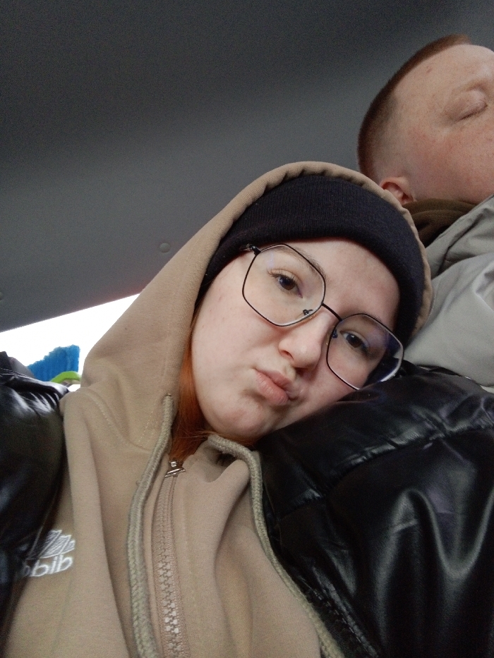 An indoor, close-up photograph depicts two individuals inside what appears to be a vehicle. In the foreground, a young individual with fair skin, reddish hair, wearing a dark knit beanie, large black-rimmed glasses, and a beige zippered hoodie over a black puffy jacket, rests their head on the shoulder of a second individual. The subject's lips are pursed, and their eyes are directed towards the camera. A partially visible white logo resembling "adidas" is present on the left chest of the beige hoodie. To the right and slightly behind, the second individual's head and shoulder are visible; they possess short, light brown hair and appear to have closed eyes, indicating rest. The background consists of a plain gray vehicle ceiling, with a blurry blue object resembling a brush or broom partially visible on the far left. The image provides no discernable information to identify the specific location within the city of Краснояр, Russia.