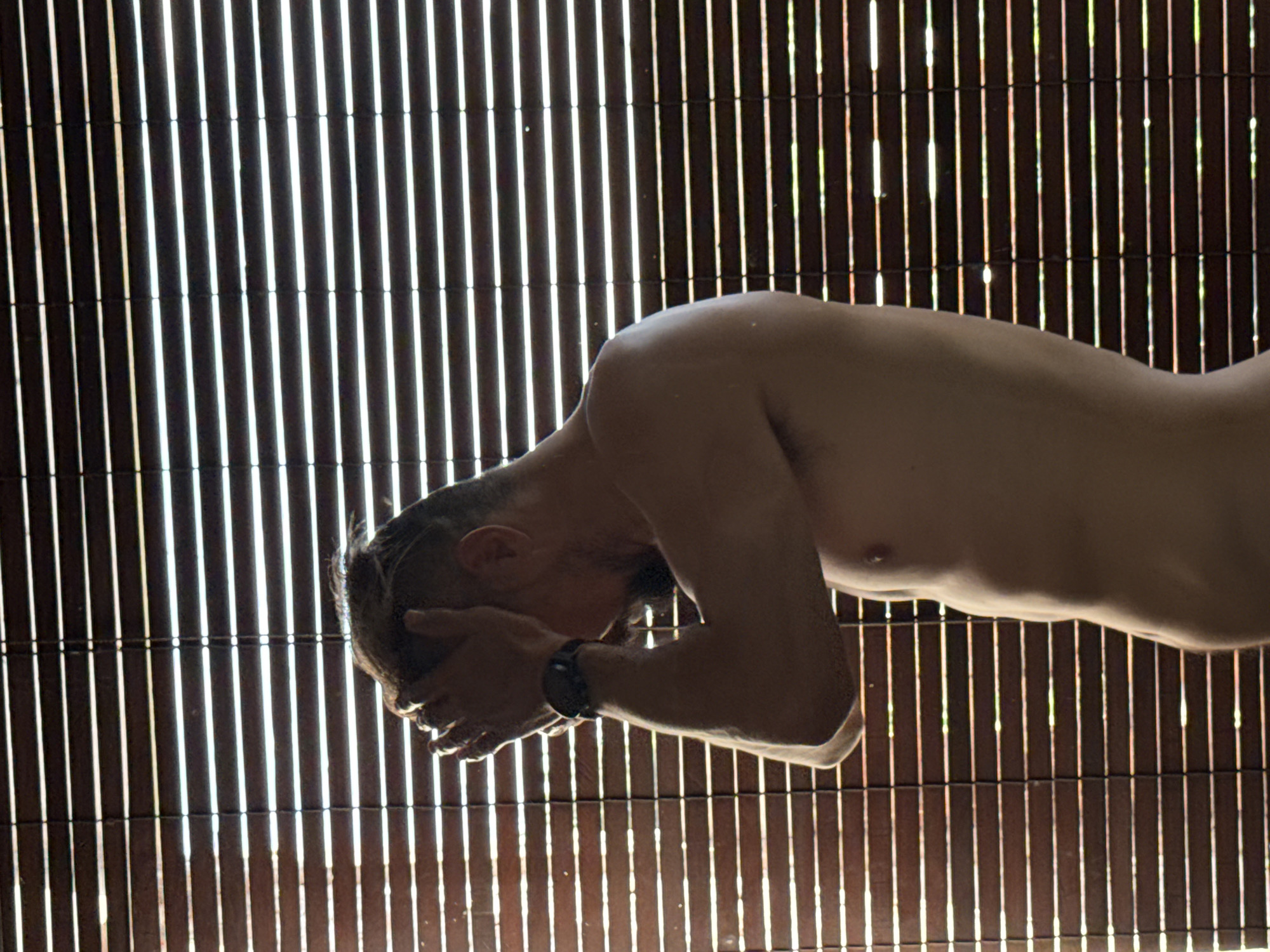An shirtless adult male stands with his head in his hands, viewed from the side, against a backdrop of horizontal blinds. The blinds are dark brown with thin strips of light filtering through, creating a striped effect. The man appears to be looking down, his face obscured by his hands. He has dark hair and a beard. A black watch is visible on his left wrist. The image appears to have been taken indoors, possibly in a bathroom or bedroom, with natural light coming from behind the blinds. The scene is likely Kuta Selatan, Indonesia, given the information provided about the location.