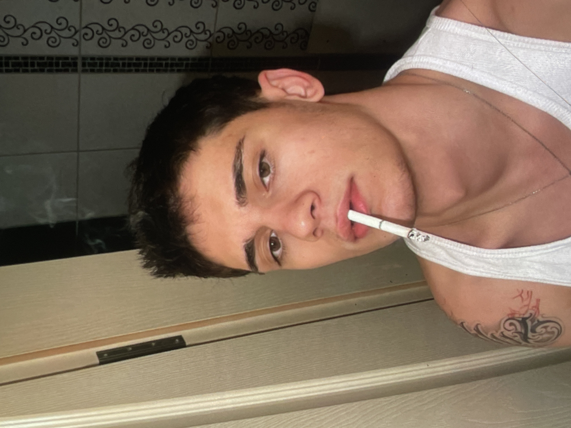 A young male with short, dark hair and brown eyes is pictured in a close-up, selfie-style shot. He is wearing a white tank top and has a cigarette in his mouth. He has a tattoo on his left shoulder and is wearing a silver necklace. The background appears to be a bathroom, with tiled walls and what looks like a doorframe to the left. The lighting is dim. The image does not provide any information about a specific location within the city of Harmanli, Bulgaria.