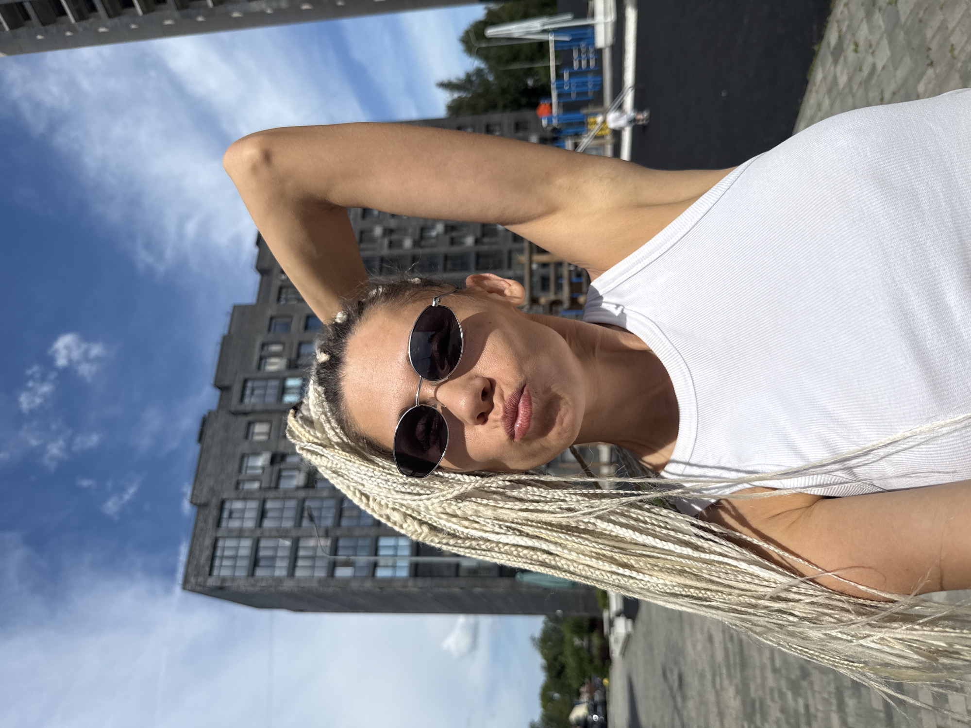 A young woman with blonde braided hair and oval sunglasses is taking a selfie outdoors. She is wearing a white ribbed tank top and has her right arm raised, with her hand resting on the back of her head. She is making a pouting face. In the background, a modern brick apartment building is visible under a blue sky with scattered clouds. To the right of the building, a section of a playground with blue and orange structures is discernible. The ground in front of her is paved with grey interlocking stones. The scene is set in the city of Rusanovo, Russia.