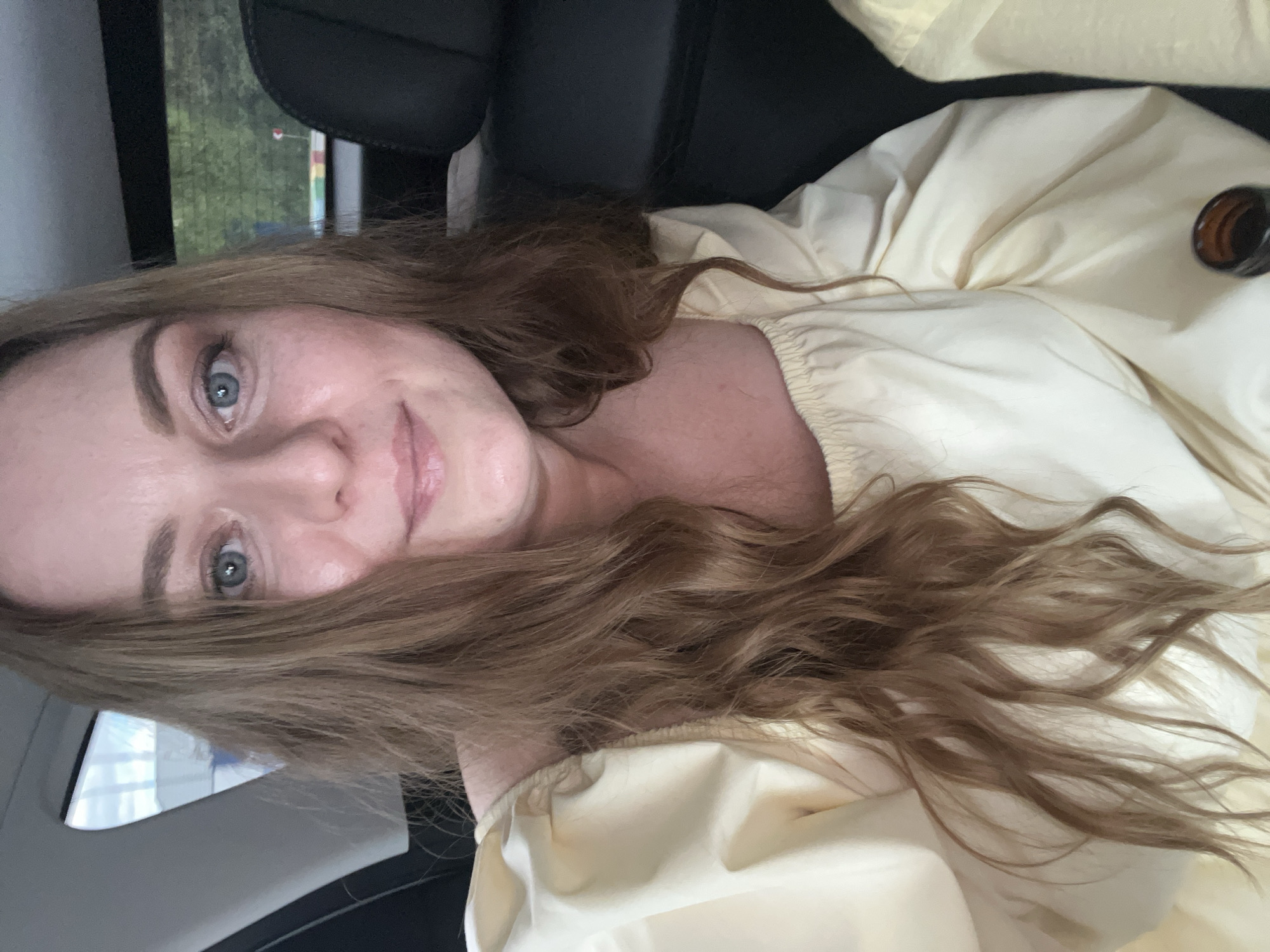 A fair-skinned woman with long brown hair and blue eyes is photographed from a close, slightly angled perspective. She is wearing a light yellow blouse. The background indicates she is likely inside a vehicle, with a car seat and window visible. The location is Nikol'skoye, Russia, although this is not discernible directly from the image. A small brown bottle is visible in the upper right corner of the frame.