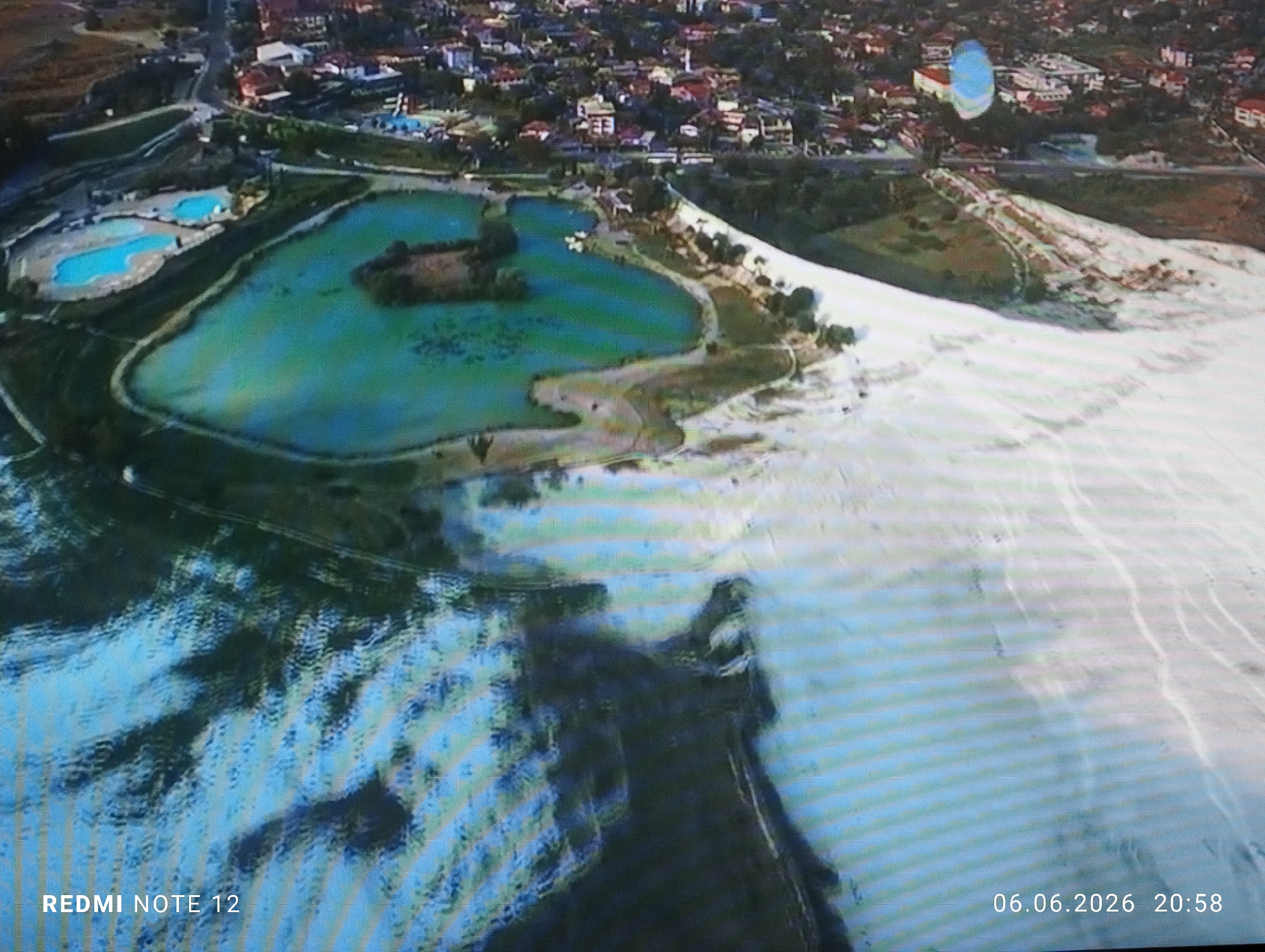 This is a low-resolution aerial image depicting a section of the Burgas Salt Pans and Lake Atanasovsko, situated within the city of Burgas, Bulgaria. The image exhibits noticeable horizontal scan lines or compression artifacts throughout. A timestamp "06.06.2026 20:58" is visible in the lower right corner, and "REDMI NOTE 12" is present in the lower left corner.
The dominant feature in the right and central portions of the image is an expansive, bright white landscape, characteristic of salt deposits or active salt evaporation beds. This white area displays undulating textures and patterns, with some darker, possibly wetter, sections interspersed, particularly at its edges where it meets land with sparse vegetation.
To the left of the white salt beds, a large body of water with varying hues of blue and green occupies a significant part of the midground. Within this body of water, several smaller, distinctly turquoise-blue pools are visible, delineated by narrow, dark green landforms, indicating controlled salt evaporation ponds. A small, irregularly shaped island covered in dark vegetation is present within one of the larger water bodies. Adjacent to these pools, further to the upper left, additional blue-toned pools are present, appearing to be outdoor swimming pools.
Beyond the salt pans and green areas in the upper background, an urban environment is visible, comprising numerous buildings with a mix of light-colored and reddish roofs, nestled among trees. Roads and paths traverse the landscape, connecting different sections of the area.
In the upper right section of the image, a single blue and white parachute or paraglider is observed in the sky, indicating a person engaged in an aerial sport. No individuals are distinctly identifiable within the scene.
The immediate foreground features choppy, dark blue water, showing ripples and reflections, indicating movement or a lower altitude perspective over a water surface. The lighting conditions suggest late evening or dusk, aligning with the recorded time.