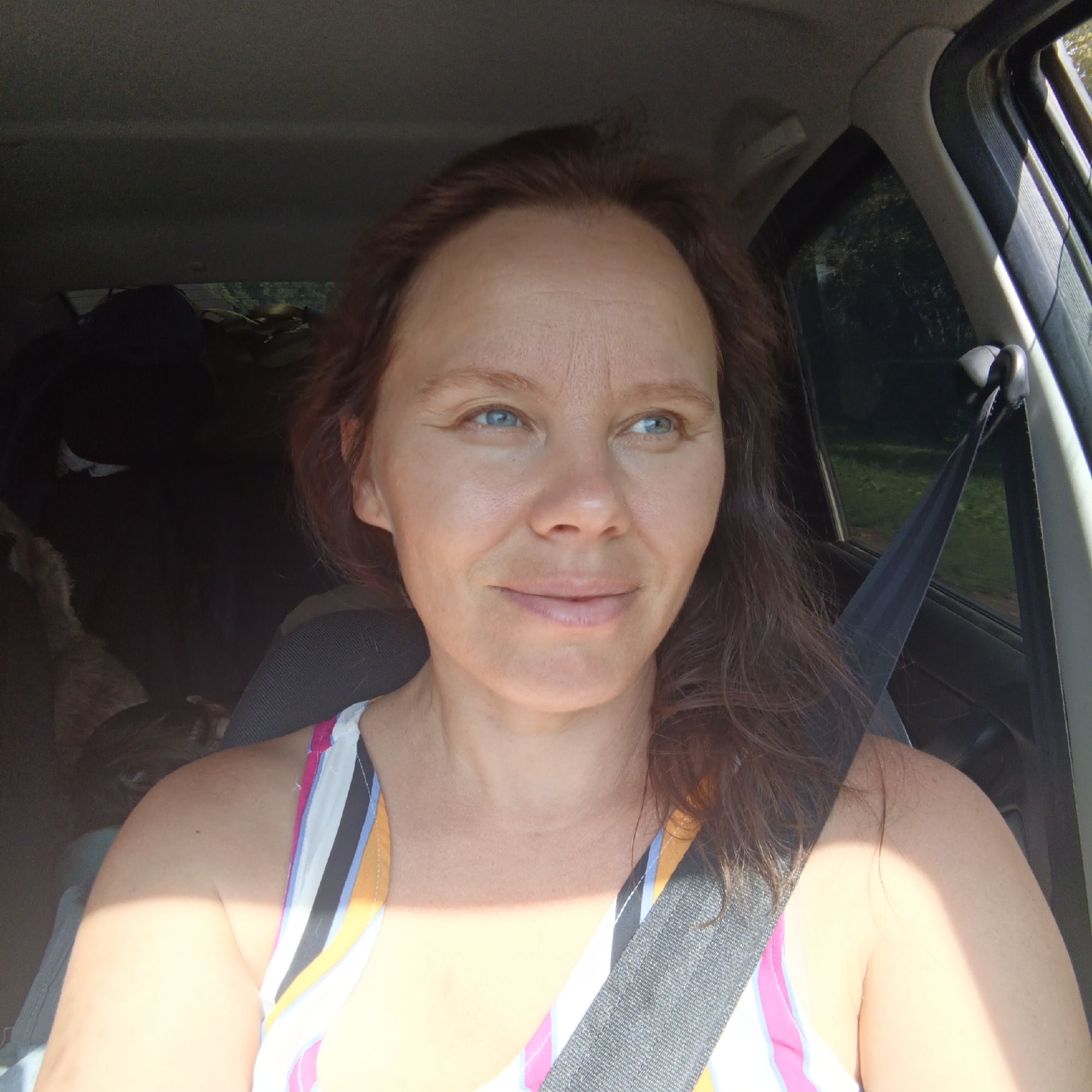 The image shows a woman in the passenger seat of a car. She has long, brown hair and blue eyes, and is looking to her right. She is wearing a colorful striped tank top and a black seatbelt. Sunlight streams in from the right, casting a shadow across her face and shoulder. In the background, the interior of the car is visible, including seats, a window with trees outside, and what appears to be luggage. The image was taken in Chishmy, Russia.