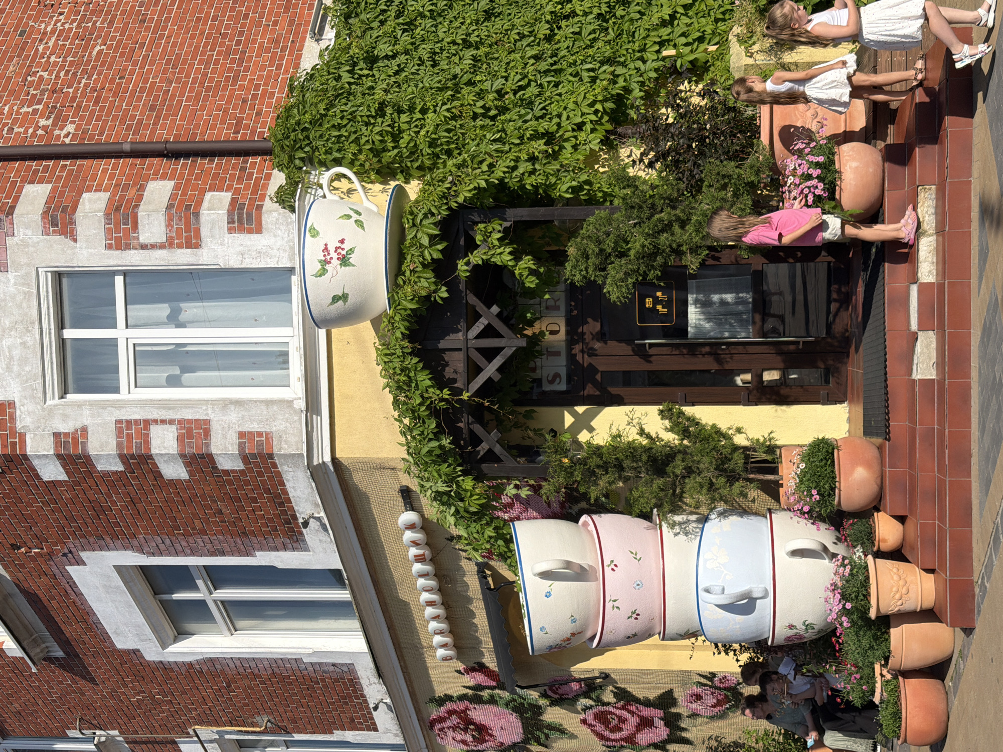 The image depicts the facade of a building in Zelenogradsk, Russia, featuring a whimsical design inspired by tea cups. Several oversized, decorative tea cups are stacked vertically in front of the entrance. The building itself is constructed with red brick and has white trim around its windows. Lush green ivy climbs the side of the building, partially obscuring the upper levels.
In the foreground, several potted plants, including those with pink flowers, are arranged on steps leading up to the entrance. Three young girls, dressed in casual summer attire, are present on the steps and sidewalk. One girl in a pink t-shirt and white shorts stands on the steps, looking towards the right. Another girl in a white dress is also on the steps, facing away from the camera. A third girl, also in a white dress, walks along the sidewalk on the right side of the image, looking down at her phone. A woman and two children are seated on a bench to the left, with the woman looking towards the children.
Above the entrance, a sign with Cyrillic letters is mounted on the wall. The entrance itself is a dark-colored door with glass panels. A small sign near the door displays operating hours. The overall scene suggests a charming and inviting establishment, possibly a cafe or shop, designed to attract attention with its unique decorative elements.