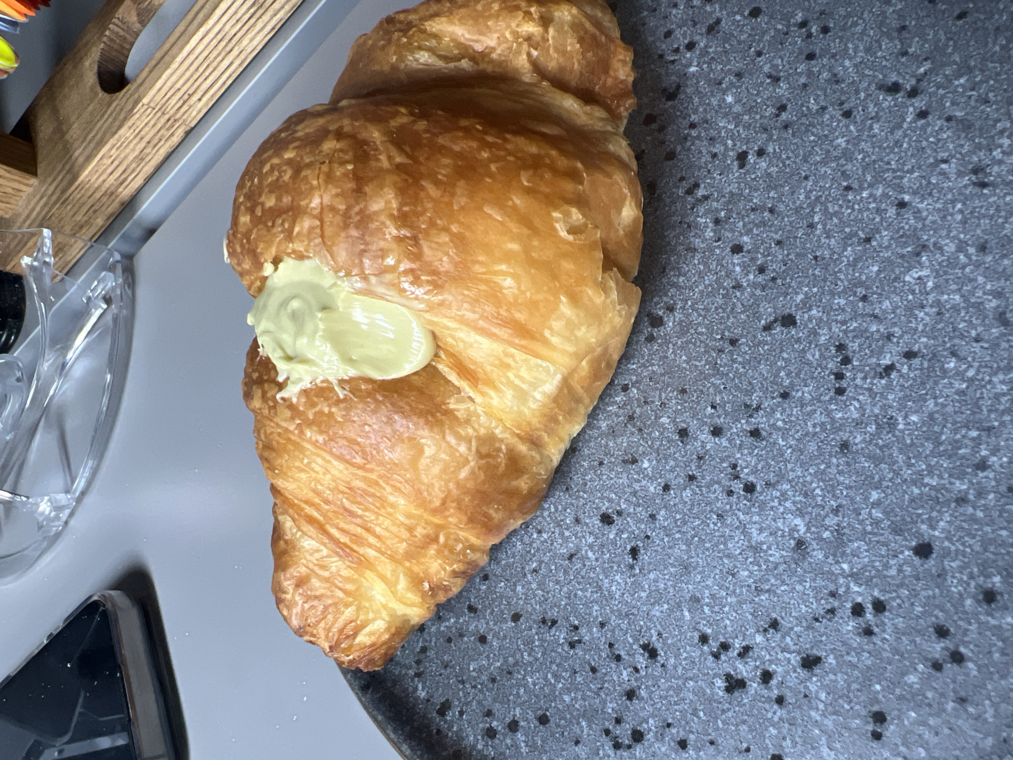 The image presents a close-up, top-down view of a golden-brown croissant resting on a speckled gray surface. The croissant is the central subject, positioned diagonally across the frame, showcasing its flaky layers and glossy, baked exterior. A generous amount of light green, creamy filling, possibly pistachio, matcha, or avocado cream, is visibly extruded onto the top center of the pastry.
In the upper left background, a wooden object with distinct horizontal slats is partially visible, suggesting a tray or part of kitchen furniture. Below this, a clear, somewhat ornate glass or plastic container is seen. The surface beneath the croissant is a dark gray material with numerous small black and lighter speckles, resembling a stone countertop or a decorative plate. A dark, possibly black, object with subtle texture is visible in the bottom-left corner.
The scene depicts an indoor setting, likely a kitchen or a cafe, focused on the prepared food item. There is no visible activity or people, suggesting a still-life presentation. The lighting does not indicate a specific time of day. No text is visible in the image. While the provided context states the location is Tbilisi, Georgia, there are no visual cues within the image itself to confirm this.