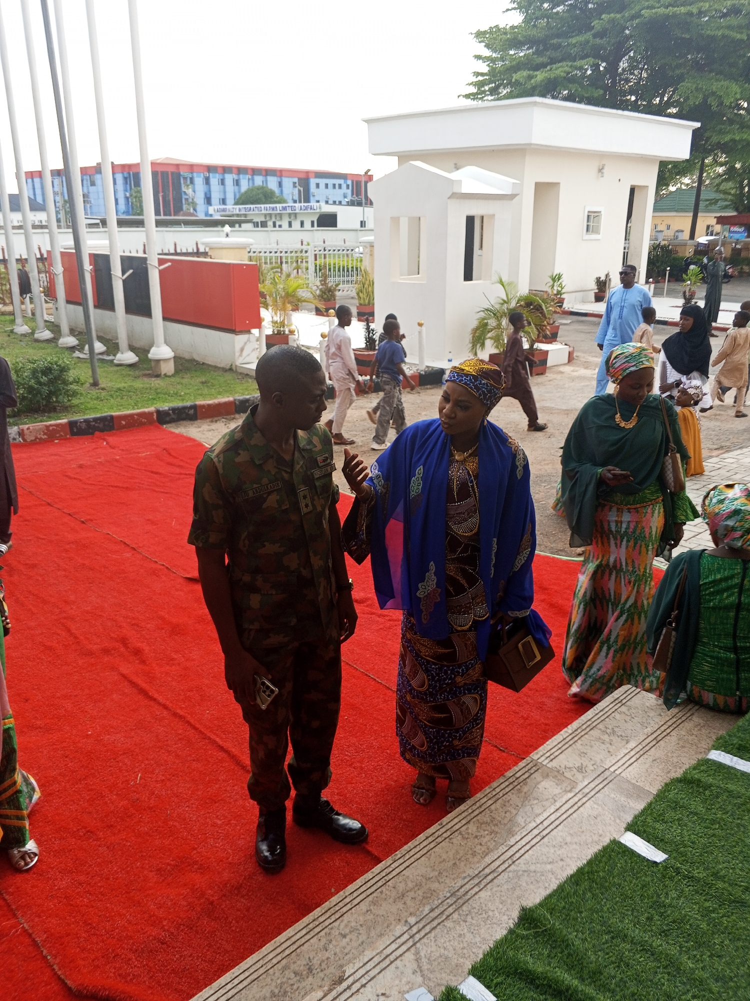 In Abuja, Nigeria, a Nigerian Army officer in camouflage uniform stands on a red carpet, facing a woman dressed in a blue kaftan and headscarf. The officer, identified by a name tag as "MAIDUN ABDULKADIR NIGERIA," holds a mobile phone. The woman, adorned with jewelry, gestures with her left hand. Behind them, other individuals in traditional attire are visible, including a woman in a green headscarf and patterned skirt. The background features a white building with a modern, cubicle-like structure and a larger building with a facade of blue and red glass panels, possibly an office complex. Trees and flagpoles are also present in the environment. The scene appears to be an outdoor event or reception, likely in a formal setting.