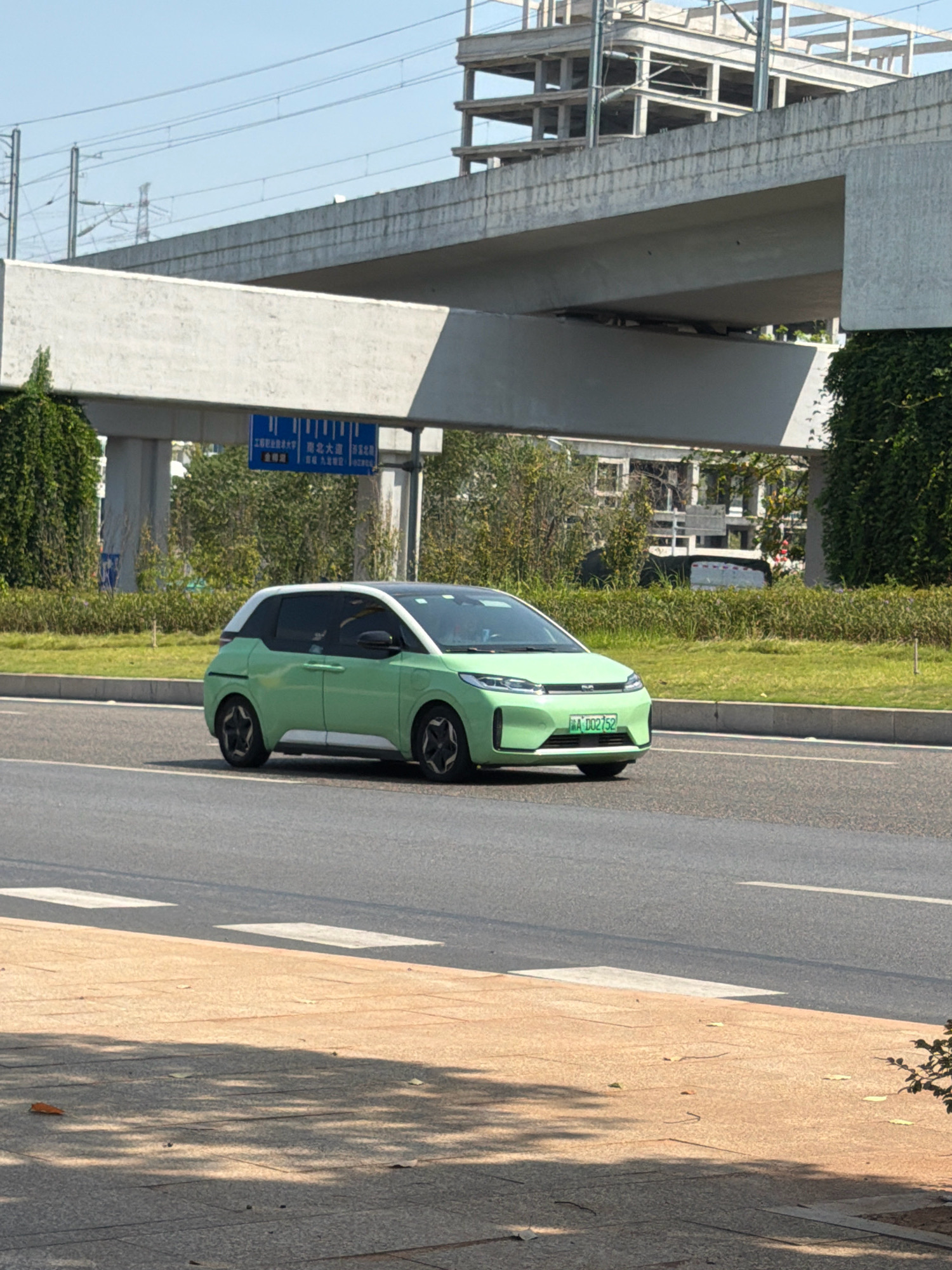 A light green electric vehicle, possibly a small hatchback, is driving on an asphalt road. The car has a white roof and black window trim, and its license plate is visible and reads "A 0D2752". The road has white lane markings. In the background, there is a concrete overpass, a partially constructed building, and trees and greenery. A blue traffic sign with Chinese characters is visible on a pole near the overpass. The scene appears to be an urban or suburban environment in Jiangjin, China, under a clear sky. The foreground shows a sidewalk or pedestrian area with shadows cast by trees.