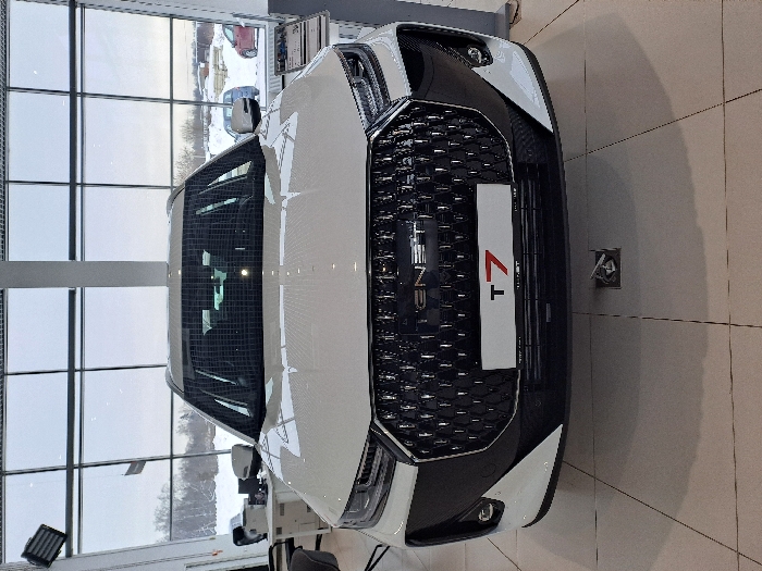 A white car, possibly an Oshan Z7, faces the camera head-on. Its black grill dominates the view, displaying the vehicle's name and model. The car is situated in a well-lit indoor space with a large window showcasing a snowy outdoor scene. The floor is covered with square tiles, and a portion of the building's ceiling is visible.