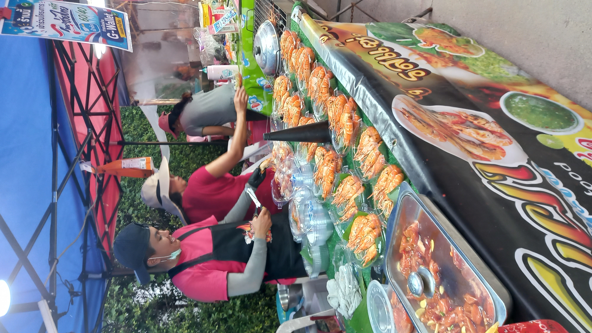 An outdoor food stall is depicted under a blue and red canopy in ตำบลเกาะพะงัน, Thailand. In the foreground, a person wearing a pink shirt, black apron, grey arm sleeves, and a black baseball cap is actively using metal tongs to handle cooked, orange-hued prawns, which are individually packaged in clear plastic containers with some resting on banana leaves. A surgical face mask hangs from the left ear of this individual. The prawns are displayed on a table covered with a green mat, suggesting artificial grass. To the left, stacks of empty clear plastic containers and a metal bowl on a white plastic chair are visible. A large metal tray with a lid containing raw marinated meat with garlic in sauce sits on the table's left side.
Behind the primary worker, another individual in a red shirt and a wide-brimmed straw hat is partially visible, also working at the stall. Further back and to the right, a third person in a grey shirt and a pink cap is visible. A grill with a metal lid is positioned on the right side of the main display table.
A white sign with blue text, displaying Thai script, a "G-Wallet" logo, and "60/40" (likely indicating a government co-payment scheme), hangs from the canopy. The front of the display table is adorned with a banner featuring colorful graphics, including images of grilled prawns, a green chili-lime dipping sauce, and Thai script alongside "Seafood" in English. Green foliage serves as the background, and the ground surface is paved.