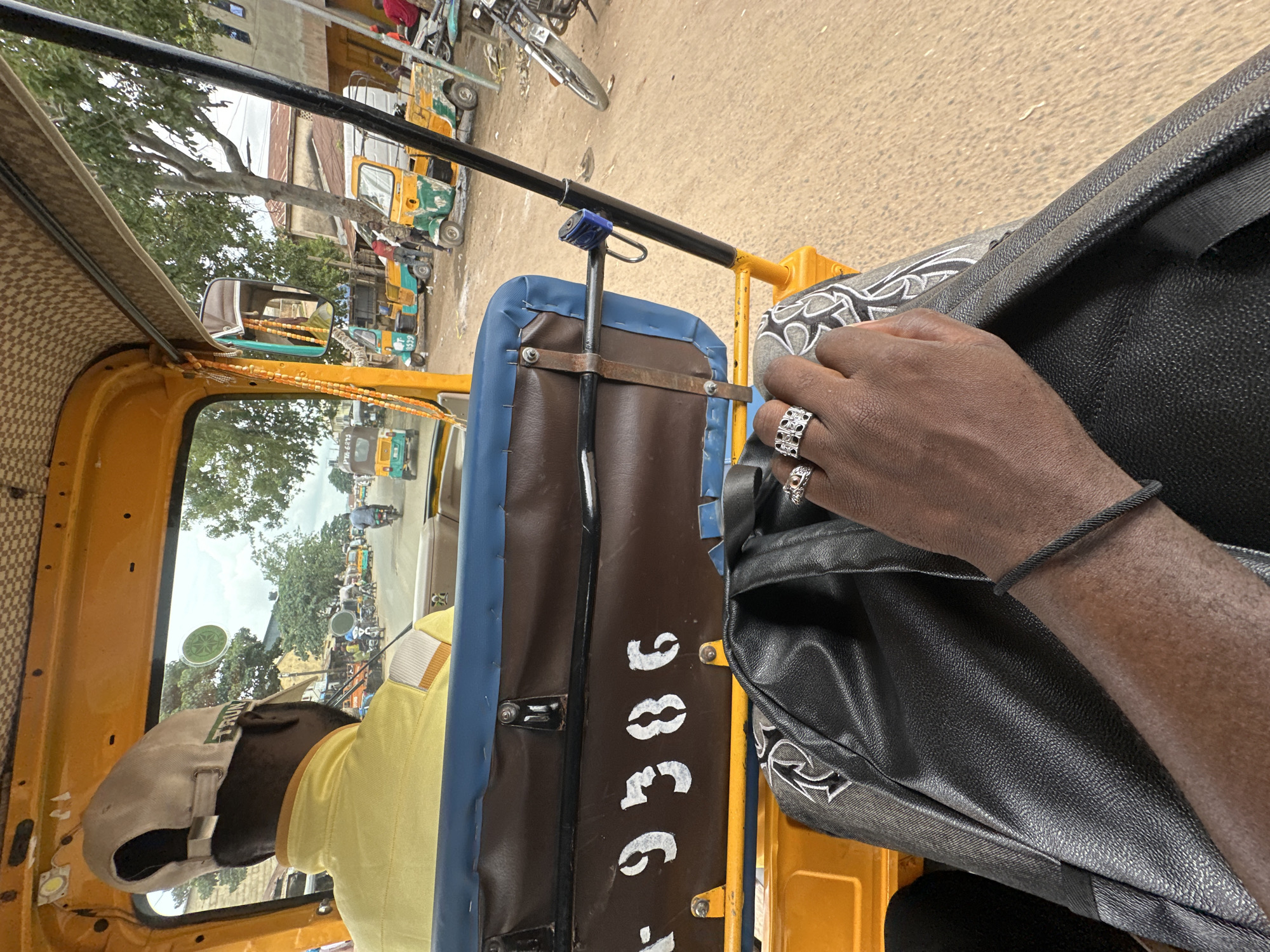 The image shows the interior of a yellow and blue auto-rickshaw, viewed from the perspective of a passenger. The driver, wearing a yellow shirt and a light-colored cap, is visible from behind, facing forward. The auto-rickshaw appears to be moving through a street in Jos, Nigeria, as suggested by the presence of other auto-rickshaws, motorcycles, and buildings in the background. There are shops and trees lining the street. The number "9386" is stenciled on the back of the passenger seat. The passenger's hand, adorned with rings, rests on a black bag on their lap.