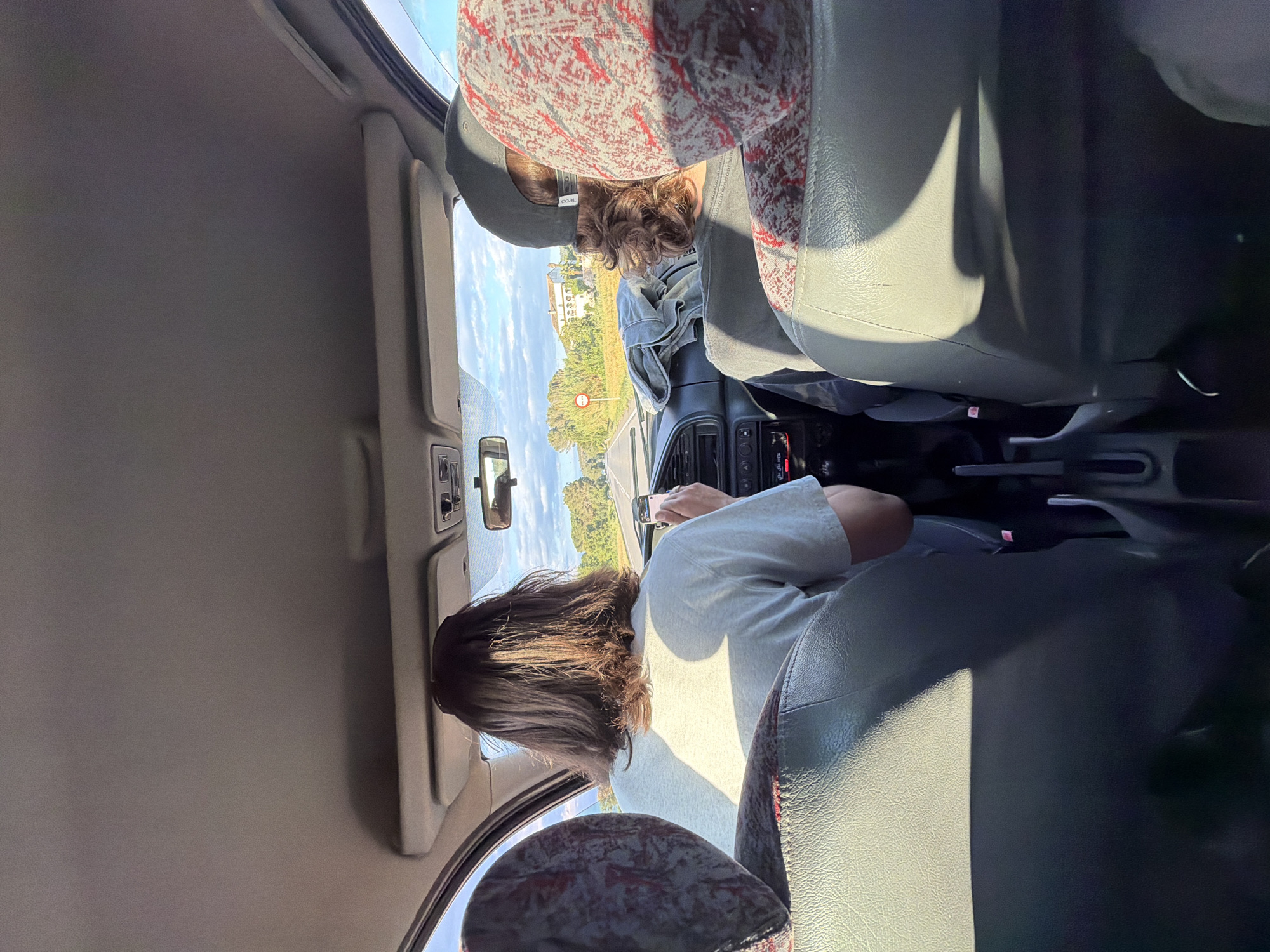 The image shows an interior car view. A person is driving while looking at a phone. Another person is in the back. Outside the car window, there is a road and green trees. The location is Torrent, Spain.