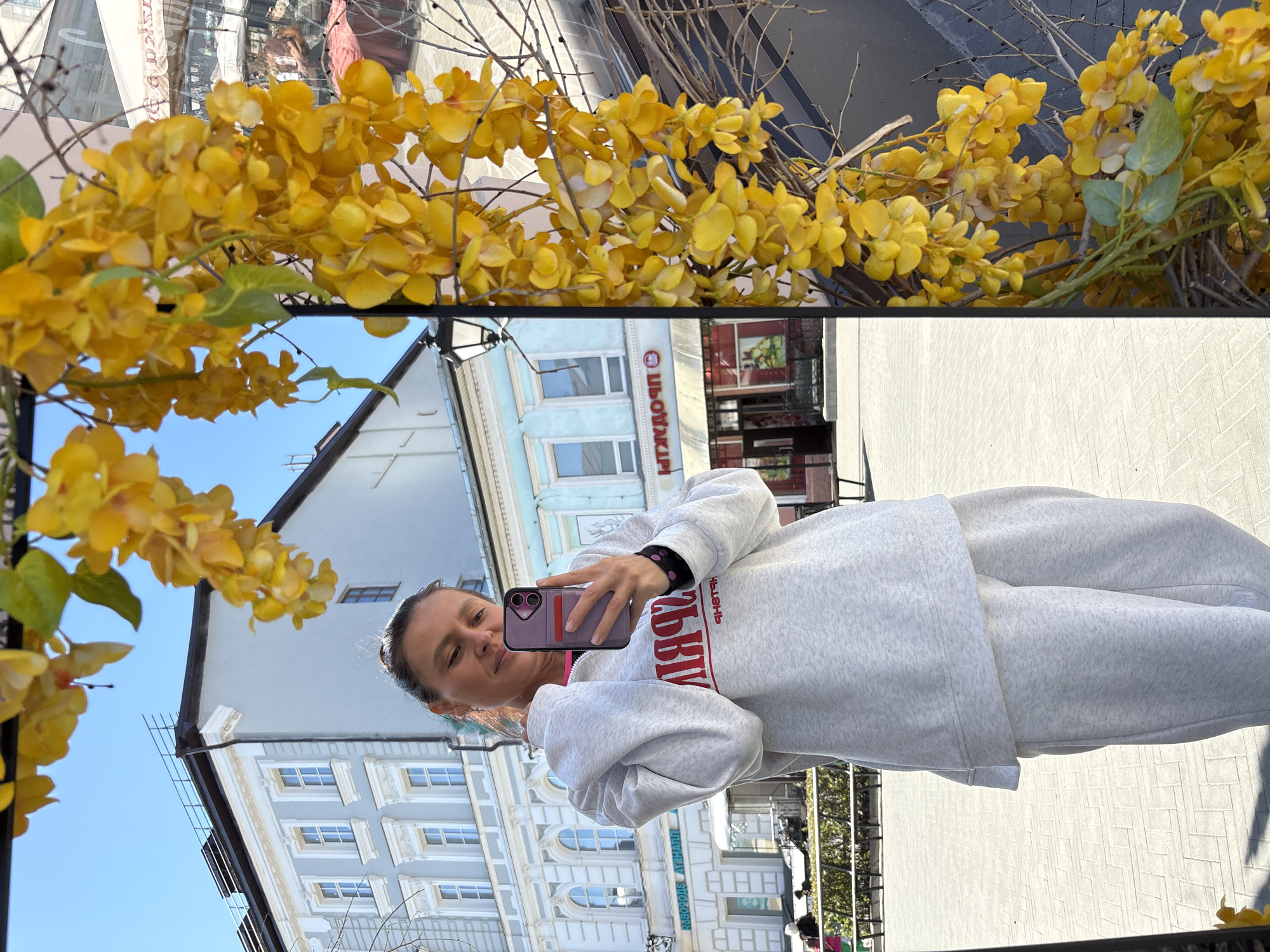 A woman is reflected in a mirror adorned with yellow flowers in Kazan, Russia. The mirror's reflection includes the woman holding a phone, wearing a grey sweatsuit, and standing in front of a building with a light blue facade. The building has multiple windows. In the background, the reflected street scene includes storefronts and pedestrians. The sky is clear. The ground is paved with brick.