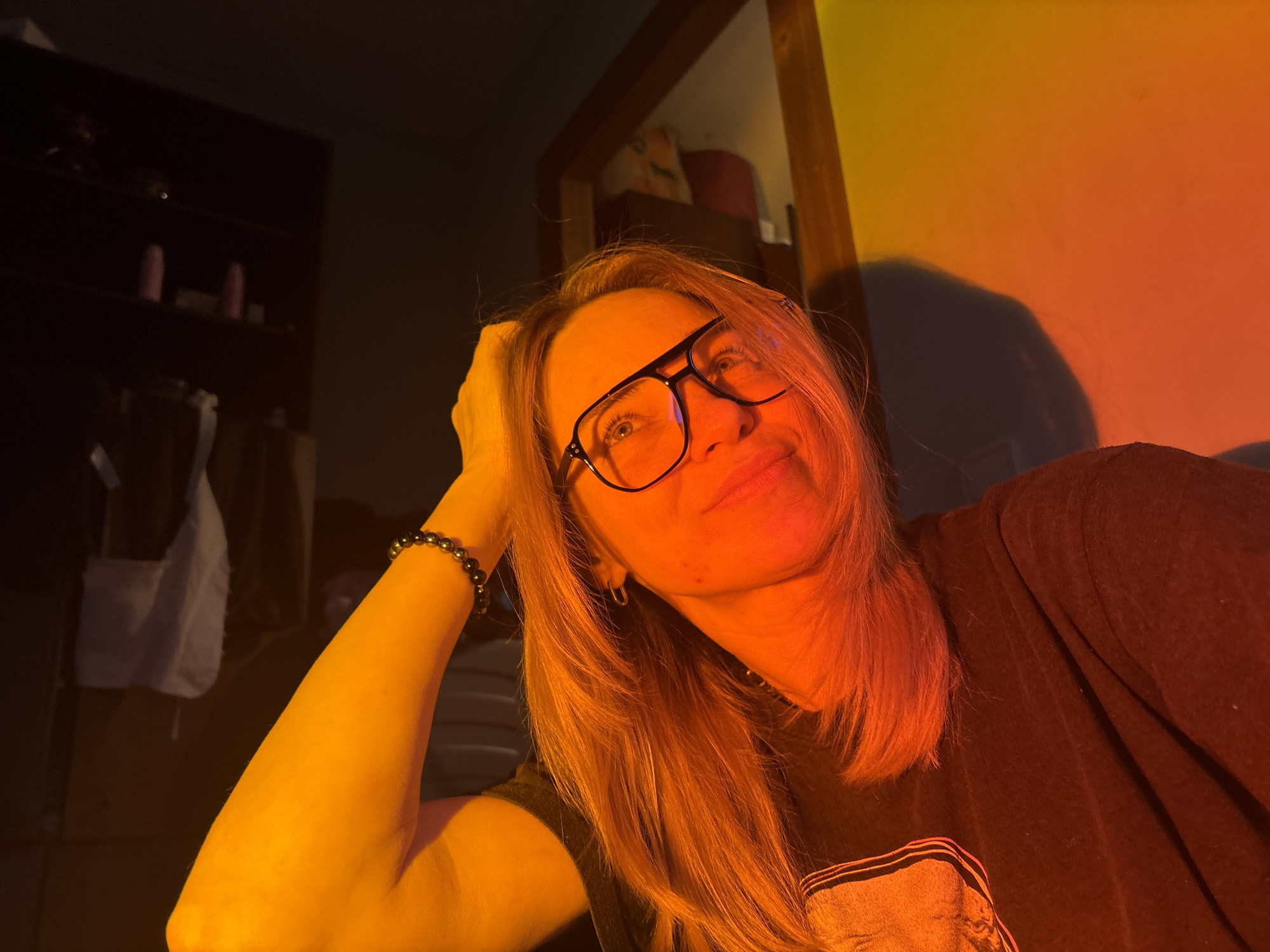 A close-up, indoor portrait captures a woman illuminated by strong, artificial colored lighting that creates a gradient from yellow to orange and red across the scene. The subject is a woman with long, light-colored hair, wearing black-framed glasses. Her right arm, adorned with a dark, beaded bracelet, is bent with her hand resting near her head. She is looking upwards and slightly to the viewer's left with a gentle, relaxed smile, suggesting a contemplative or content mood. Her body is angled slightly, and she appears to be wearing a dark t-shirt, parts of which are visible on the right and bottom left of the frame.
The background on the left features a dark shelving unit or cabinet, where some indistinct objects, possibly bottles, are visible, along with a white bag hanging. On the right, a wooden doorframe or trim is visible, and the wall behind the woman displays the strong orange and red light, casting a soft, dark shadow of her head and shoulders. The overall setting appears to be a cozy, personal space, possibly a room at home. There is no discernible text visible in the image. The unique lighting creates a warm and intimate atmosphere.