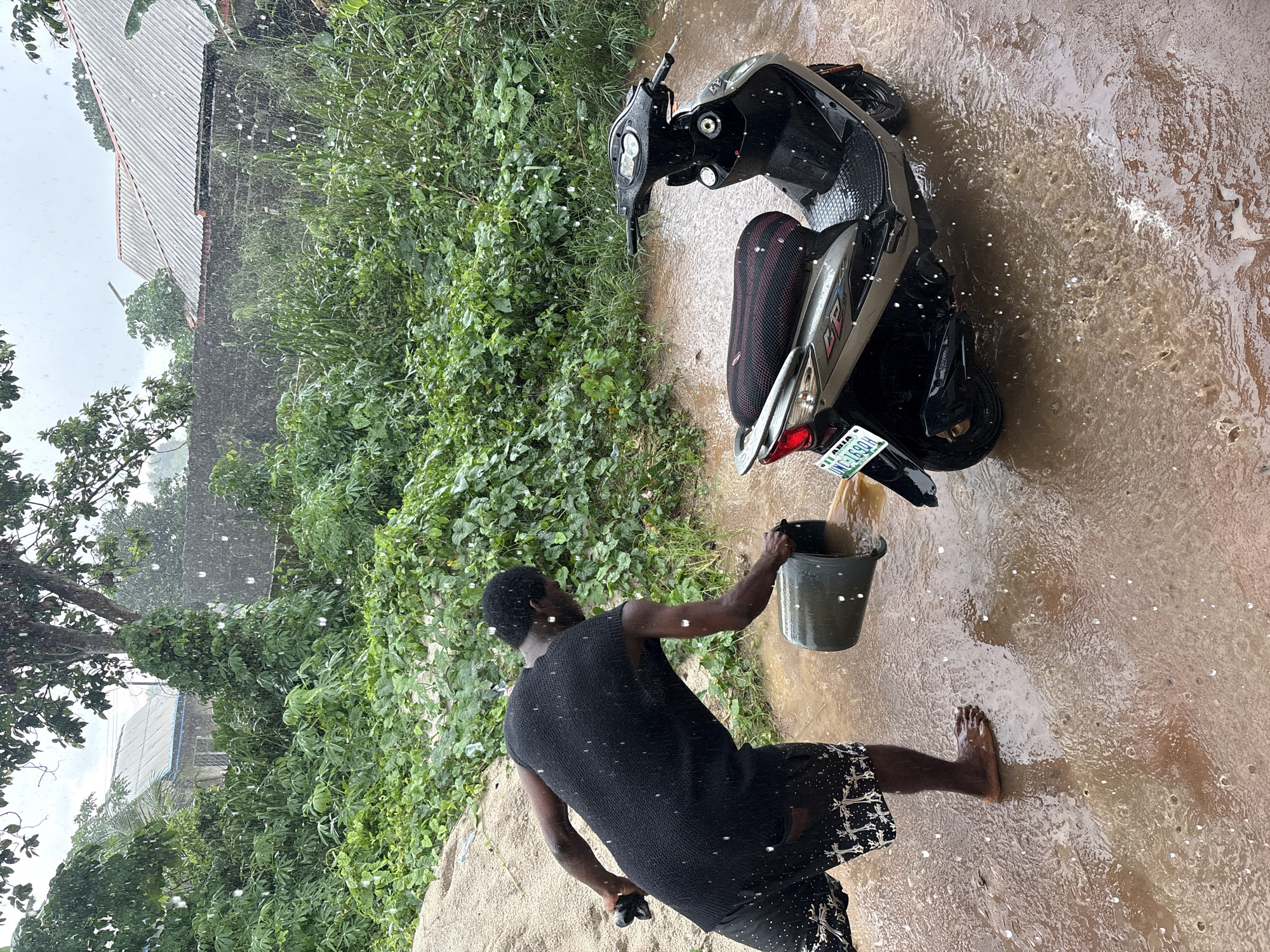 A man is shown outdoors washing a motorcycle in a rainstorm, likely in Ikot Ekpene, Nigeria based on the license plate, which reads "UNC-169DH, ASIA". The motorcycle is mostly dark with a red and black seat. The man is dark-skinned and barefoot, wearing a sleeveless black top and patterned dark shorts. He is holding a gray bucket and pouring water over the motorcycle. The ground is covered in muddy water and there is dense green vegetation behind the motorcycle. A building with a corrugated metal roof is visible in the background. It is raining heavily, with many visible raindrops throughout the scene.