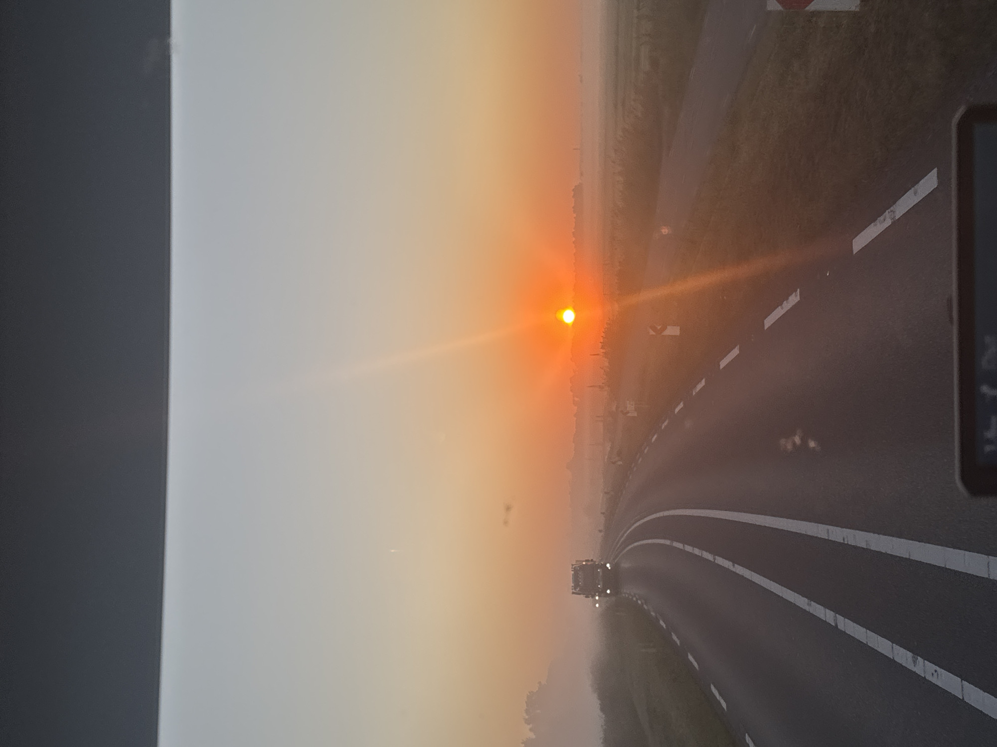 The image captures a sunrise over a road in Lekkerkerk, Netherlands. A truck is driving away from the viewer on the wet asphalt road, which is marked with white lines. The sun, a bright orange orb, is low in the sky, casting a warm glow and creating lens flare. Mist or fog hangs low over the fields on either side of the road, partially obscuring the trees and buildings in the distance. The sky transitions from a pale blue at the top to orange and yellow near the horizon. A dark bar runs across the very top of the image.