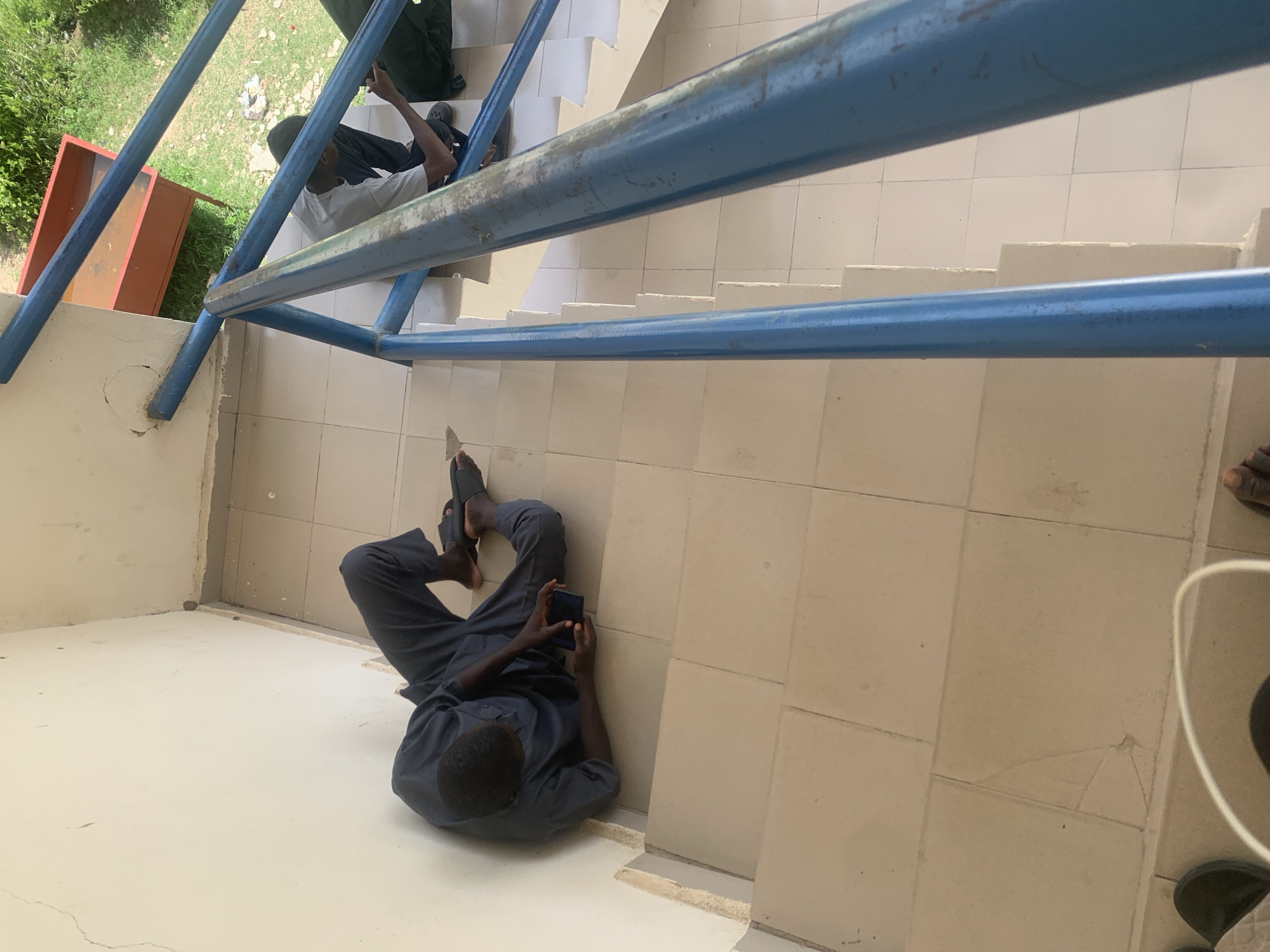 A man is seated on a stairwell, leaning against a cream-colored wall. He is wearing dark blue traditional attire and black flip-flops, and is holding a dark-colored smartphone in his hands, appearing to be engaged with it. The stairwell has tiled steps and is bordered by a blue metal railing. In the background, another person is visible further up the stairs, also seated and looking at a phone. Outside the stairwell, a red metal bin and green vegetation are visible. The scene is likely outdoors, possibly in a public space within Jalingo, Nigeria.