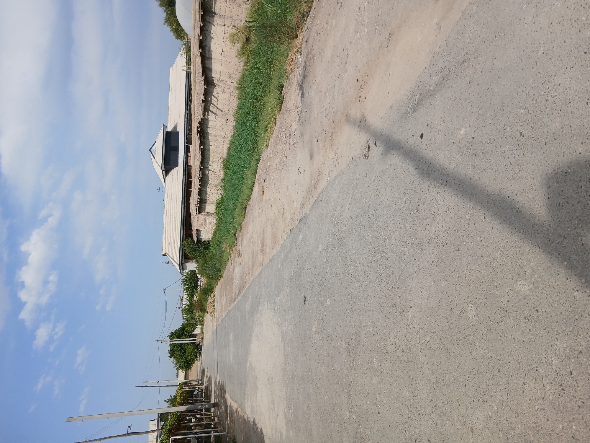 The image shows a paved road in a rural or suburban area. On the left side of the road, there are some structures with white roofs, possibly houses or agricultural buildings, with some vegetation and grapevines. On the right side of the road, there is a low, rough wall made of earth or brick, with green grass and vegetation growing along its base. Beyond the wall, there is a large building with a distinctive roof structure. The sky is blue with scattered white clouds. The overall scene suggests a warm, sunny day. There are no people or animals visible in the image. The road appears to be empty, with a shadow cast across it, likely from a utility pole or tree.