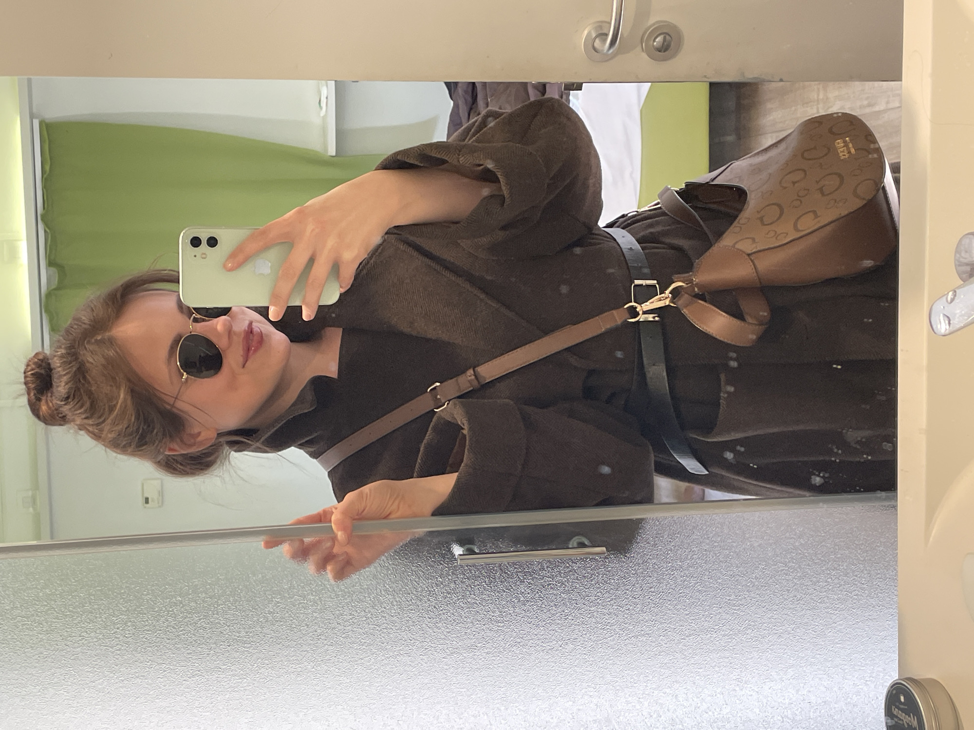 A young woman wearing sunglasses and a brown robe with a handbag stands in front of a mirror, taking a selfie with her smartphone. She is located in what appears to be a bathroom, evidenced by the shower glass in the lower portion of the frame and the bathroom fixtures (faucets, dispensers) in the background. The background also shows a green curtain. It is not possible to determine the location in the city of Venice, Italy from the image.