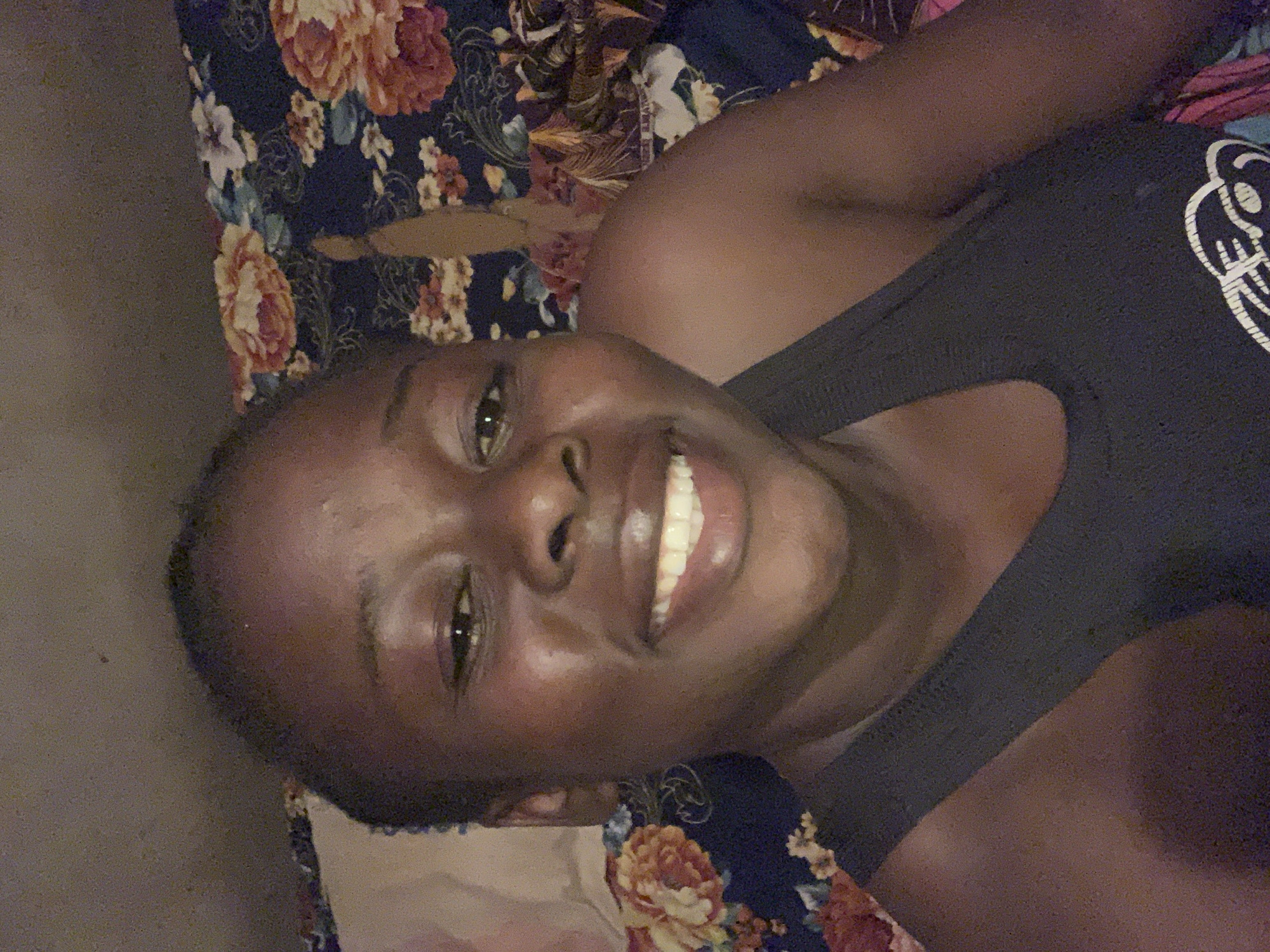 A person with dark skin and short hair is smiling and lying on a bed. The bedding has a floral pattern with blue, peach, and brown colors. The person is wearing a dark gray tank top with a white logo on the right side. The location is potentially a bedroom in a residential area; however, the city of Agban, Nigeria cannot be confirmed from the image.