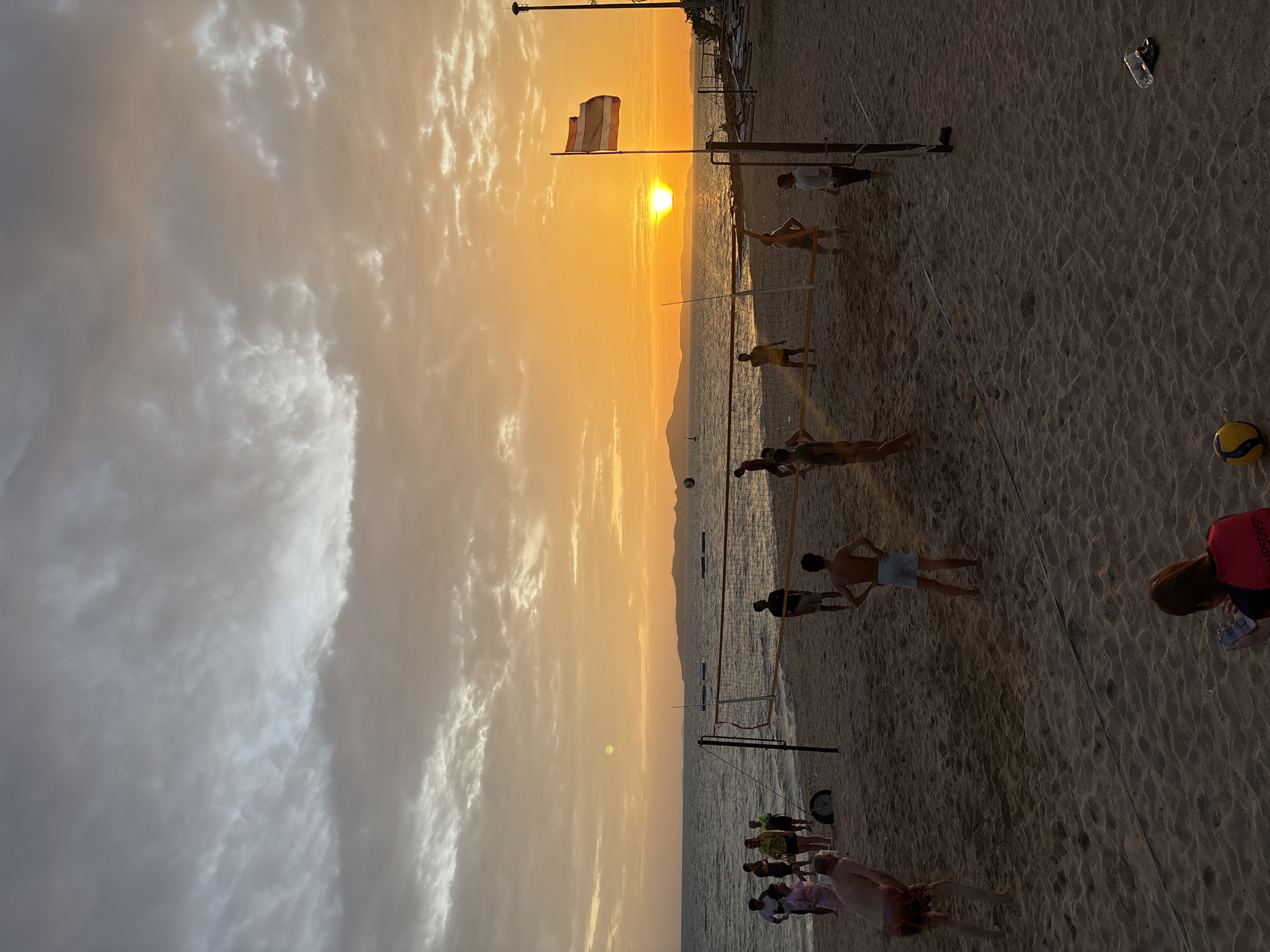 This image depicts a beach volleyball game at sunset in Pattaya City, Thailand. The sky is filled with dramatic, colorful clouds, with the sun prominently setting over the ocean. A volleyball net is set up on the sandy beach, with several individuals playing or observing the game. There are men and women on both sides of the net, engaged in playing volleyball. In the foreground, a person is seated on the sand, looking at a phone. The water of the ocean is visible in the background, with a few boats or buoys in the distance. A Thai flag is flying on a pole to the right of the frame. The overall scene is one of recreational activity and natural beauty.