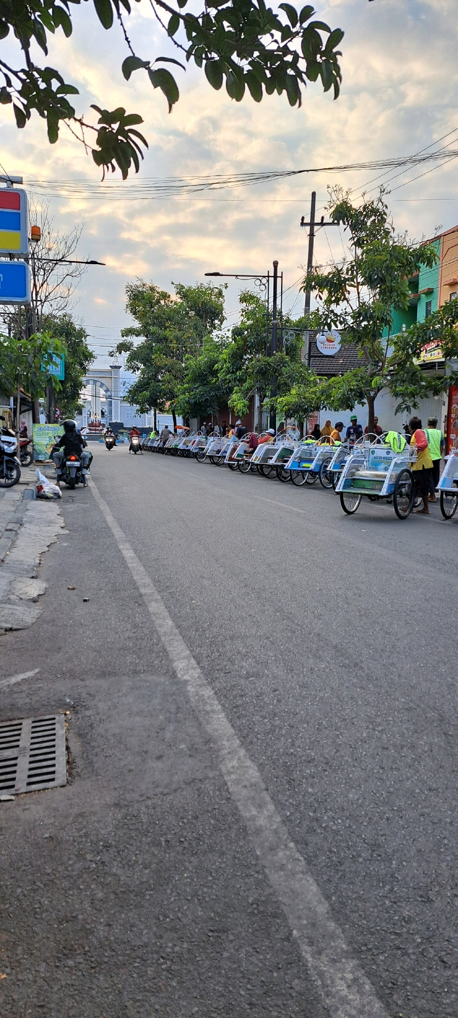 This is a ground-level, medium shot of a street in Pasuruan, Indonesia, likely in the late afternoon or early evening, as indicated by the soft light and scattered clouds in the sky. The street is lined on the right with a row of parked becaks (a type of three-wheeled passenger vehicle). Several people are standing or sitting in or around the becaks. In the background, there are trees and buildings. On the left side of the street, there is a sidewalk with a few parked motorcycles and a trash grate. Overhead, tree branches with green leaves are visible, and some power lines crisscross the sky. The overall atmosphere is one of a relatively quiet street scene in an Indonesian city, possibly a tourist area given the presence of becaks.