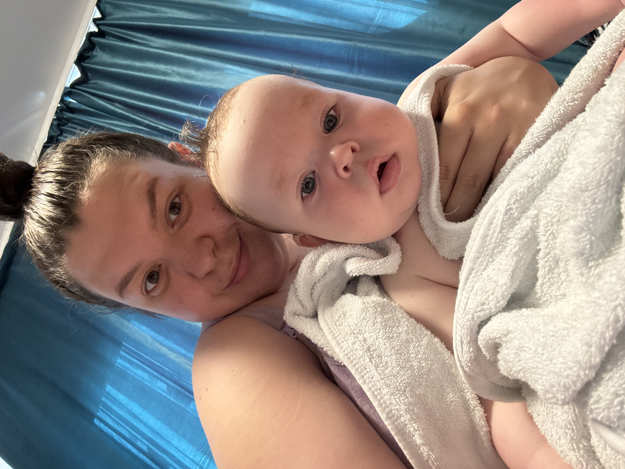 A woman with dark hair pulled back in a bun is holding a baby wrapped in a white towel. The woman is looking at the camera with a slight smile. The baby is looking at the camera with a slightly open mouth. The background is a blue curtain. The image appears to be taken indoors. The location given is Марковский, Russia, but there is no information in the image to confirm that it was taken there.