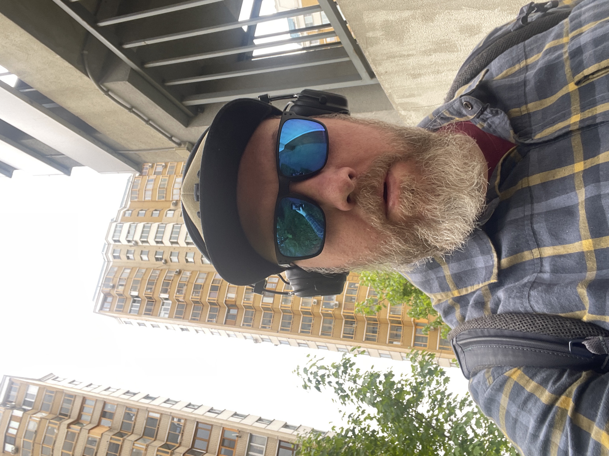 A man with a gray beard, sunglasses, a cap, and headphones is wearing a gray and yellow plaid shirt and a backpack, facing the camera from a low angle. Buildings with multiple windows are visible in the background. Based on the architecture of the buildings, this image was likely taken in the city of Khimki, Russia.