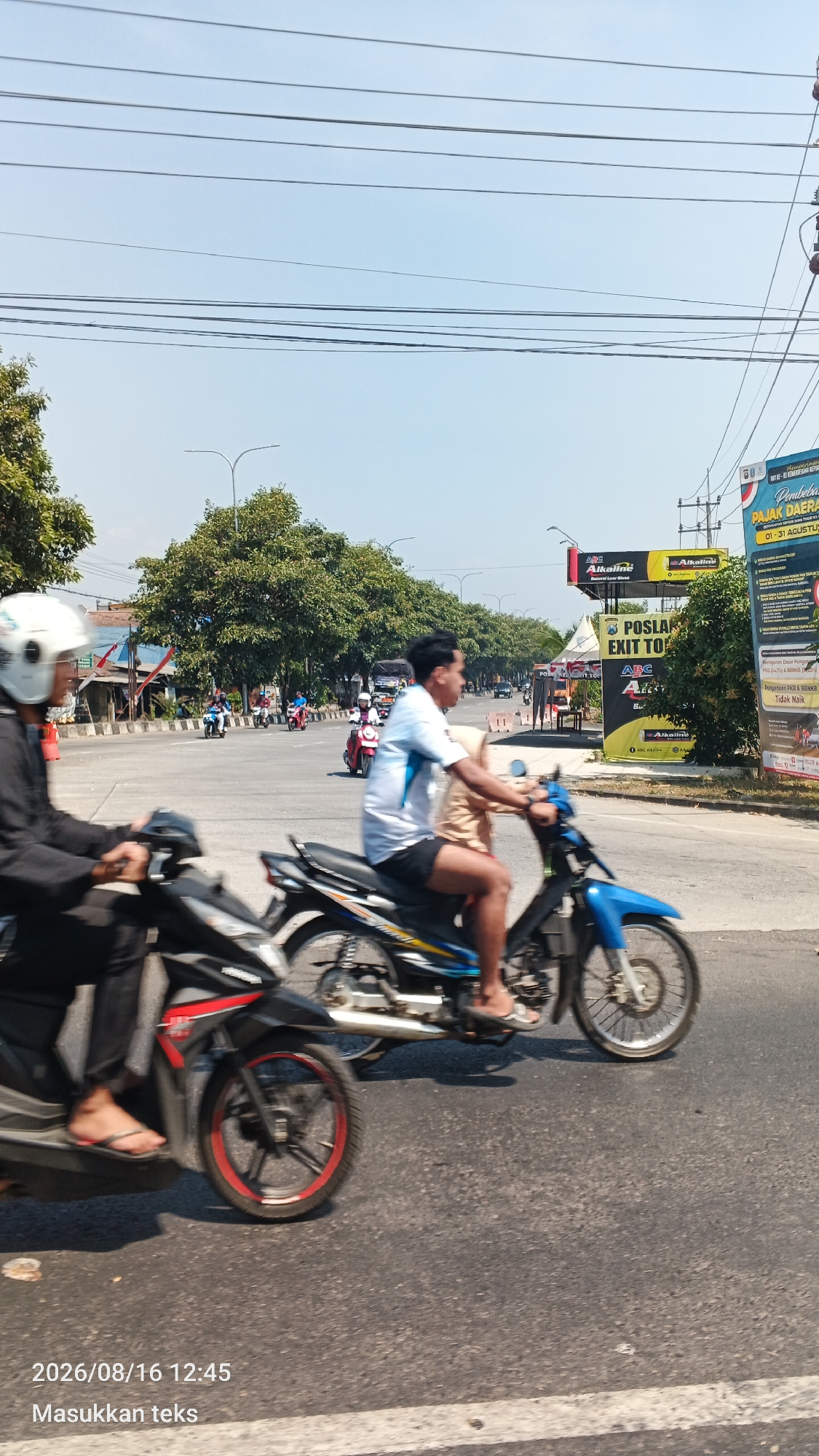 The image depicts a street scene in Bunut Wetan, Indonesia, on a clear day. The foreground features two motorcycles in motion. The motorcycle on the left is black and red, with its front wheel visible. The rider is wearing a white helmet and a dark jacket, with their feet, wearing flip-flops, resting on the footrest. The motorcycle on the right is blue with yellow and blue accents, and its rider, wearing a light blue and white t-shirt and dark shorts, is also in motion. The rider's feet, wearing flip-flops, are visible.

In the background, several other motorcycles are seen traveling on the road. The street is lined with trees and buildings, including a prominent billboard with advertisements. The sky is clear and blue, with power lines crisscrossing overhead. The overall impression is of a typical day in an Indonesian town, with ongoing traffic and local signage visible. The timestamp on the image indicates it was taken on 2026/08/16 at 12:45.