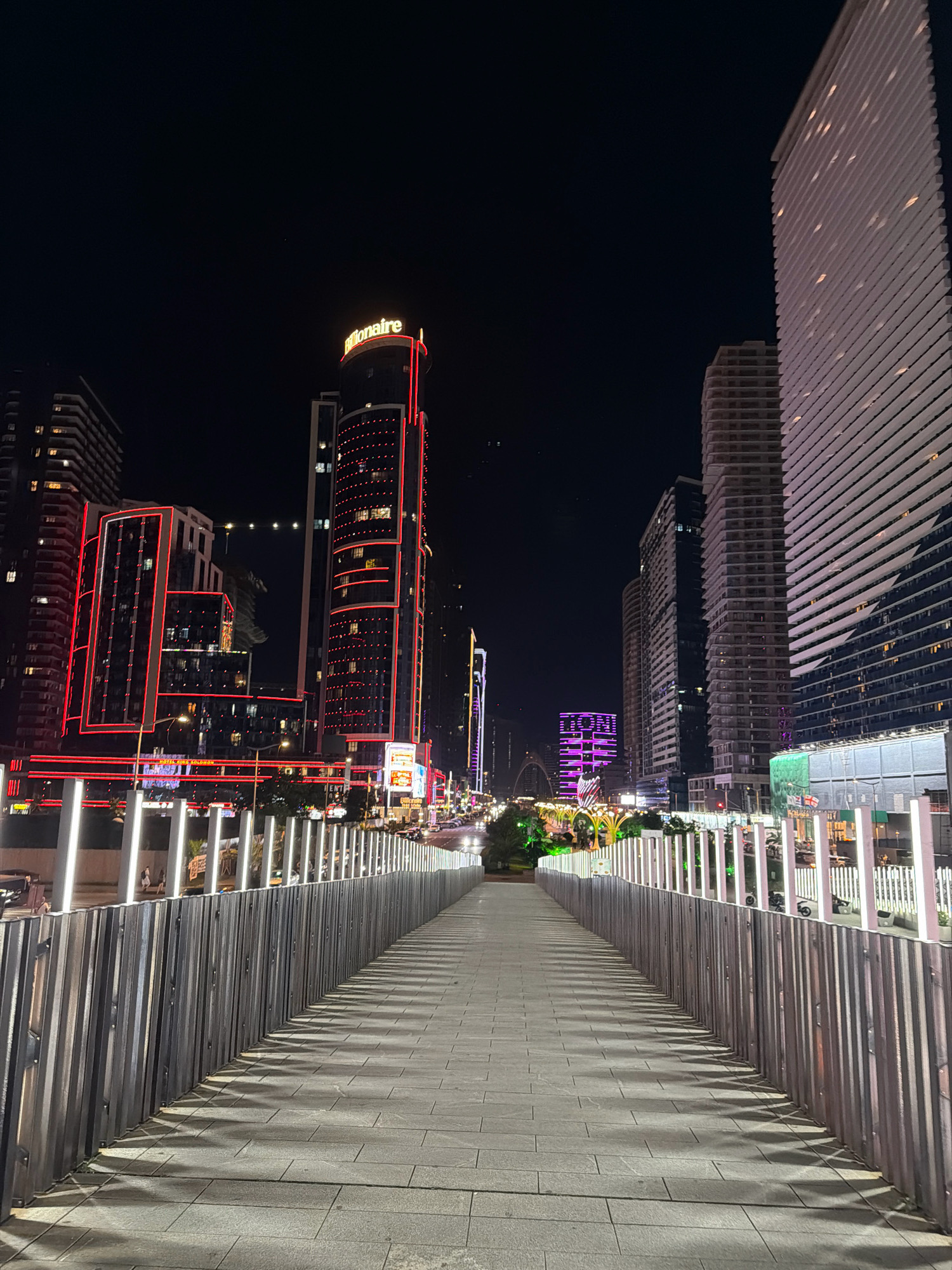 A nighttime street-level shot of a modern pedestrian bridge made of concrete with metal railings, leading into a city illuminated by numerous colorful lights. The bridge's path is made of gray cobblestone, and the railings cast long, patterned shadows onto the path, indicating a bright light source. The bridge's railing on the right is partially obscured by a metal fence. On the left side of the bridge, a row of vertical white lights line the path, casting illumination.
Beyond the bridge, a bustling cityscape unfolds. A prominent, tall, cylindrical building stands out with a red neon sign that reads "Billionaire" at its summit. This building is adorned with vertical red lights. Adjacent to it are other high-rise buildings, many of which are lit up with red and white lights, creating a vibrant and dynamic urban scene. In the distance, a building glows with purple light, and a large archway is visible. The street below the bridge is lined with cars, indicating traffic. The general area is identified as Batumi, Georgia.