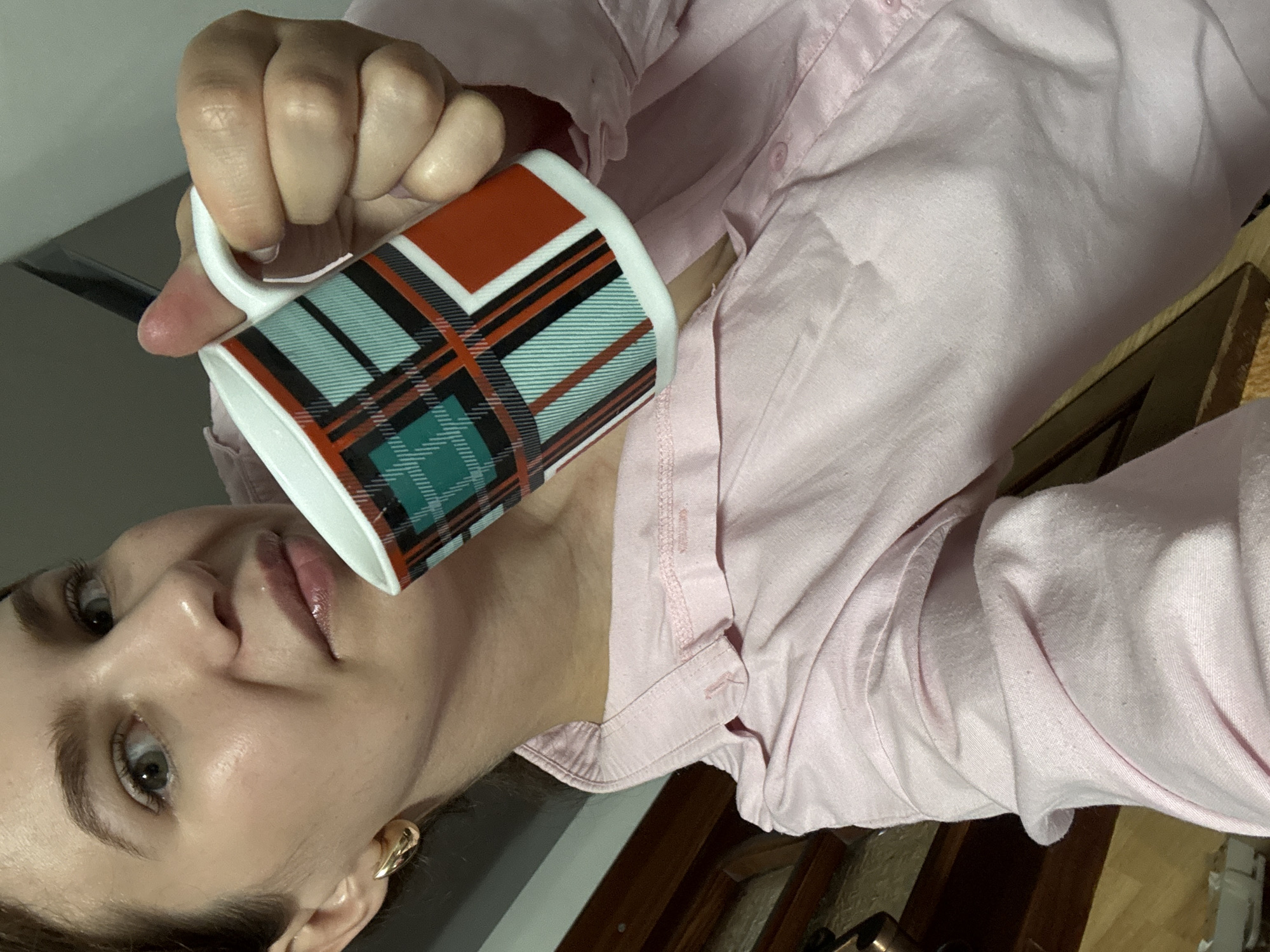 The image shows a close-up of a woman holding a plaid mug. She is wearing a pink collared shirt. The mug has a pattern of green, black, and red lines forming a plaid design. The woman has fair skin, light-colored eyes, and is wearing a gold hoop earring. The background is blurry and appears to be indoors, with hints of furniture. There is no identifiable information within the image that would suggest its location in Chaikovsky, Russia.