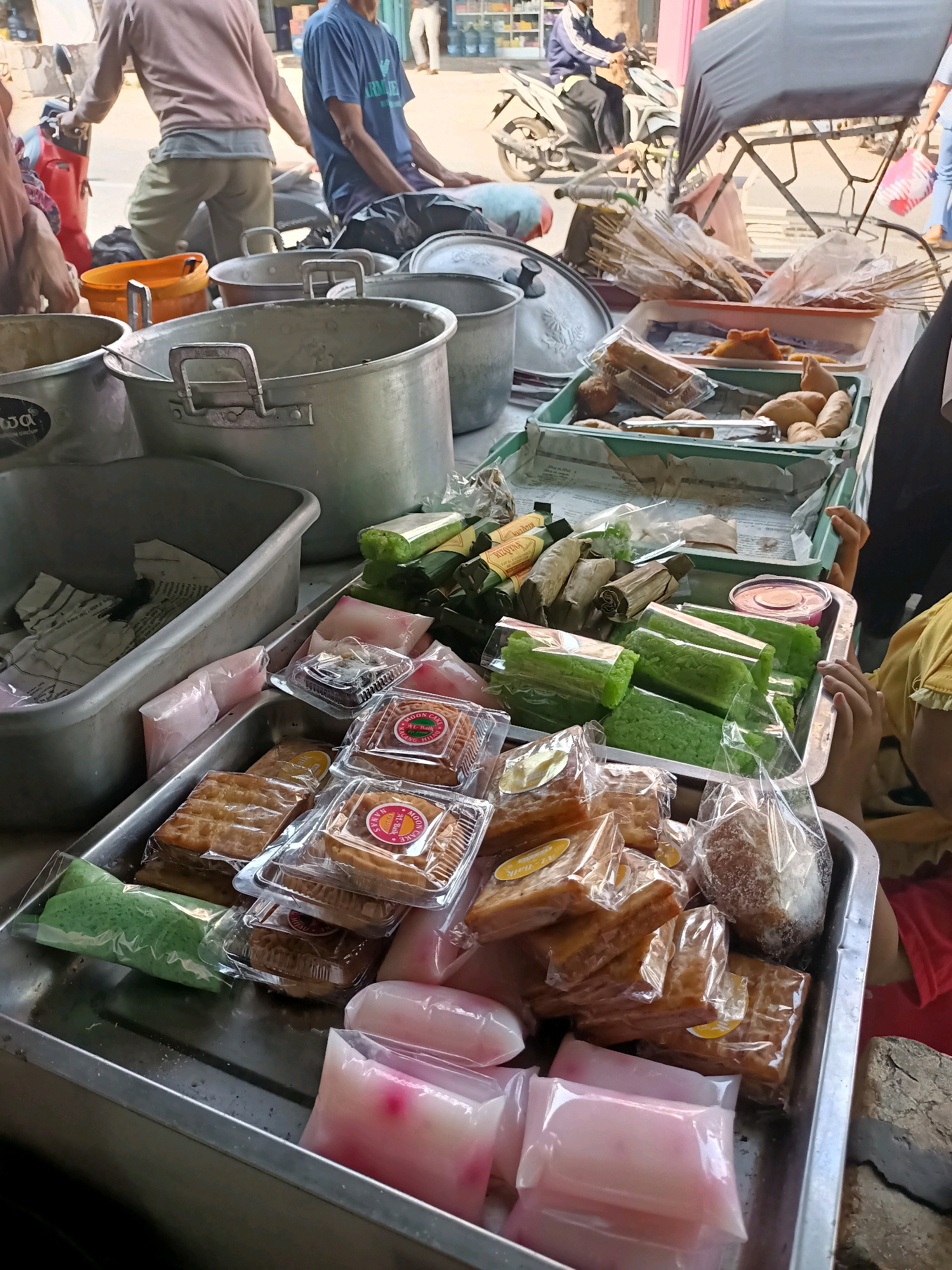 In Badas, Indonesia, an outdoor street market scene unfolds. A stall displays various packaged sweets. In the foreground, a silver tray holds an assortment of food items. To the left, green, rectangular cakes are individually wrapped in clear plastic. Next to them, small, square cakes with yellow labels are packaged in clear plastic containers. In the center and right, layered, golden-brown pastries are also in clear plastic packaging. At the bottom of the tray, several pink and white rectangular packets, likely containing a jelly-like dessert, are visible. Behind this tray, a large, aluminum pot with handles sits. Further back, other food items are displayed in green and white plastic containers, including what appear to be more wrapped pastries and green, cylindrical cakes. In the background, a person on a motorbike is visible, along with other market-goers and a shaded stall structure. The overall impression is of a bustling food vendor at a local Indonesian market.
