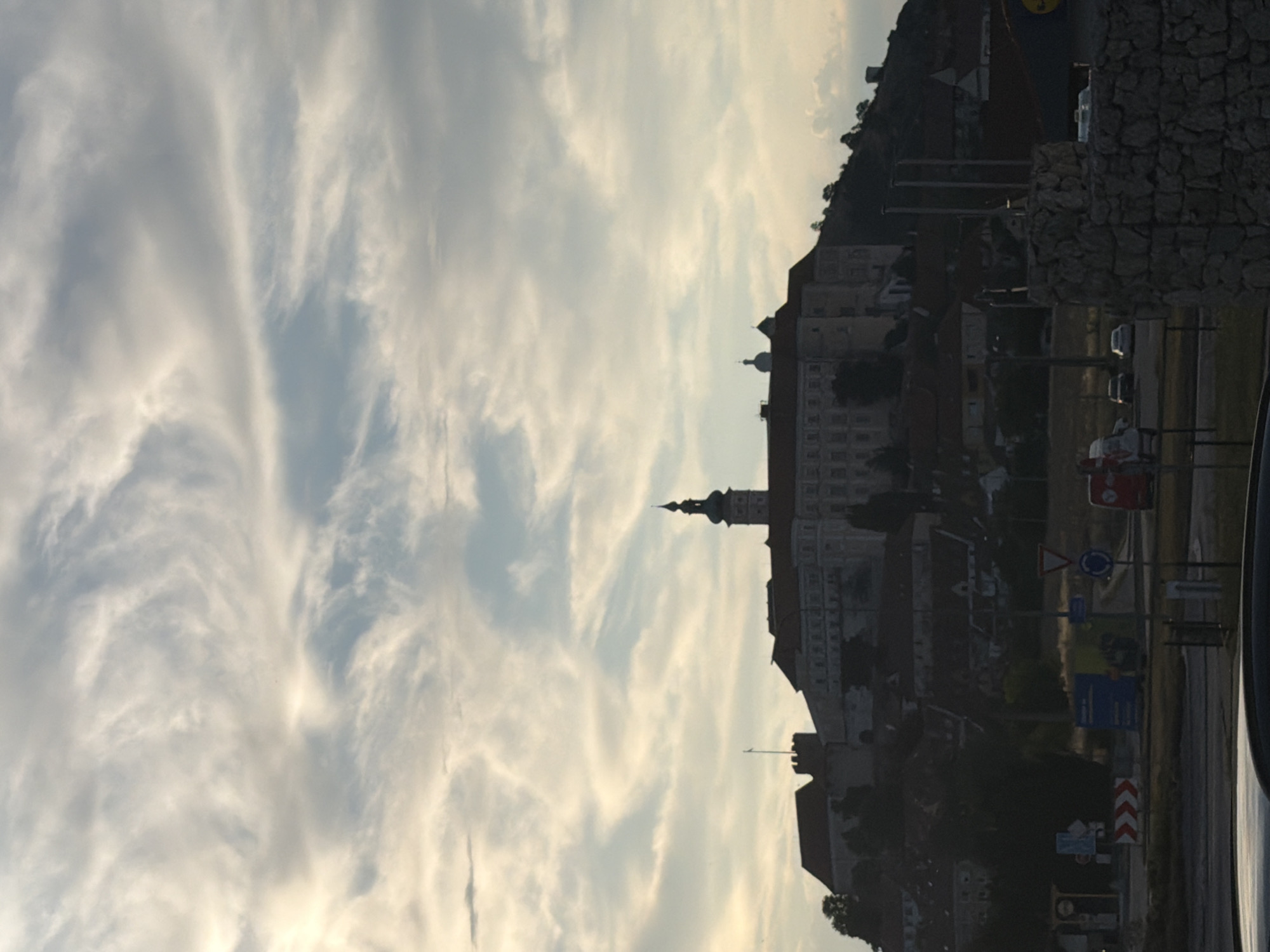 The image shows a view of Mikulov Castle in Mikulov, Czechia, from a distance. The castle, a prominent landmark, is situated on a hill overlooking the city. It is depicted in silhouette against a sky filled with wispy clouds, illuminated by the soft light of either dawn or dusk. In the foreground, there are signs of urban infrastructure, including road signs and what appear to be buildings or shops. The overall scene captures a scenic view of the historical city and its castle, with a sense of calm and atmospheric lighting.