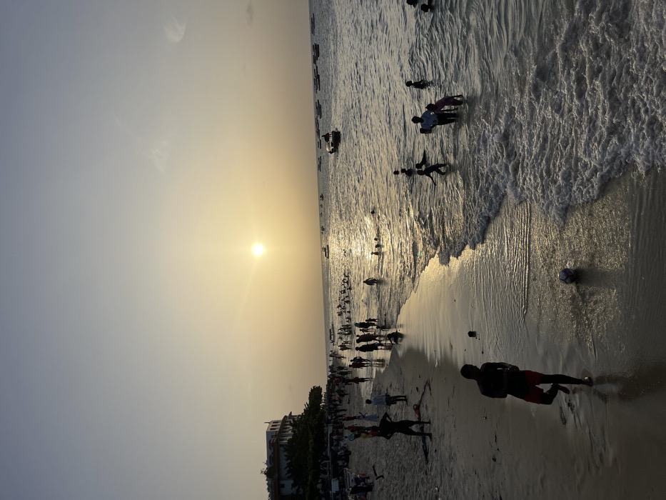 The image depicts a lively beach scene in Zanzibar, Tanzania, at sunset. The sun, a brilliant golden orb, is low on the left horizon, casting a bright reflection across the water and illuminating the hazy sky with warm hues.
Numerous people are visible across the sandy shore and wading in the ocean. Many are silhouetted against the bright light, their forms dark against the shimmering water and sky. In the foreground on the right, a person in dark shorts stands on the wet sand near the breaking waves. Other individuals and groups are spread along the beach and immersed in the shallow waters, enjoying the evening. A white soccer ball is partially visible on the wet sand near the water's edge.
Gentle waves lap at the shore, and several small boats are scattered further out in the expansive ocean. On the far right, a line of buildings and trees indicates a coastal settlement. The sky displays a soft gradient from blue to golden-orange, with subtle, wispy clouds near the top. The scene evokes a sense of communal relaxation and enjoyment of the natural beauty during the transition from day to night. There is no visible text in the image.