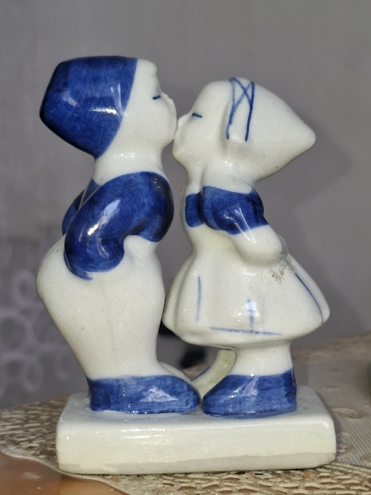 The image shows a ceramic figurine of two children kissing. The figurine is white with blue accents. The boy is on the left, wearing a blue cap and blue overalls. The girl is on the right, wearing a blue ribbon in her hair and a blue dress. They are standing on a white rectangular base. The background is a plain gray wall and a textured surface. The image is taken from a slightly elevated angle, with the figurine in the center. There is no information in the image that suggests a location in Almaty, Kazakhstan.