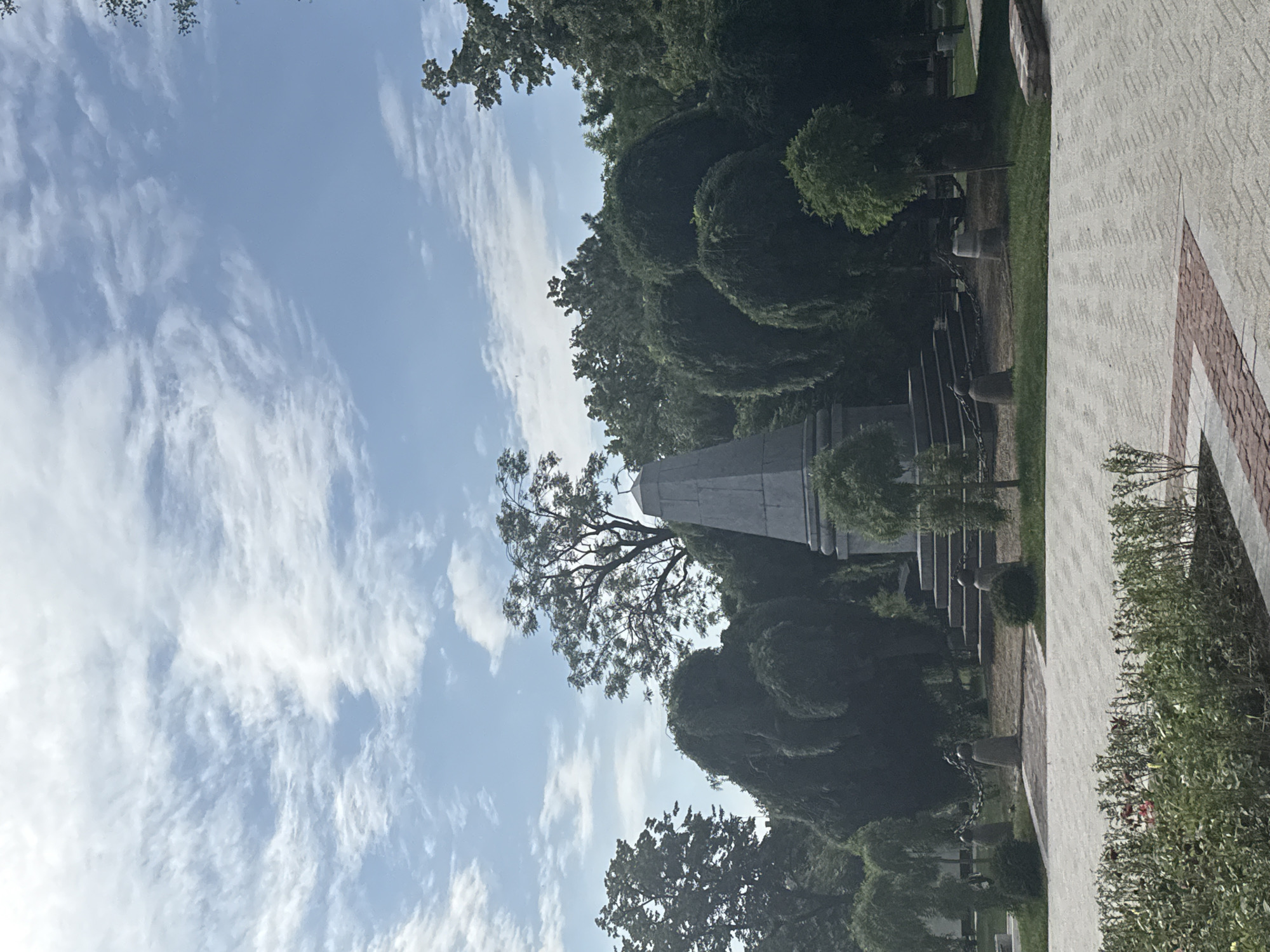 A large, stepped monument stands in a park in Stara Zagora, Bulgaria, under a partly cloudy blue sky. The monument is made of stone and tapers to a point at the top. It is surrounded by manicured trees and bushes. A paved pathway with a red and white brick border leads towards the monument, with a low hedge of rose bushes in the foreground. The park appears peaceful and well-maintained. There are no people visible in the image.