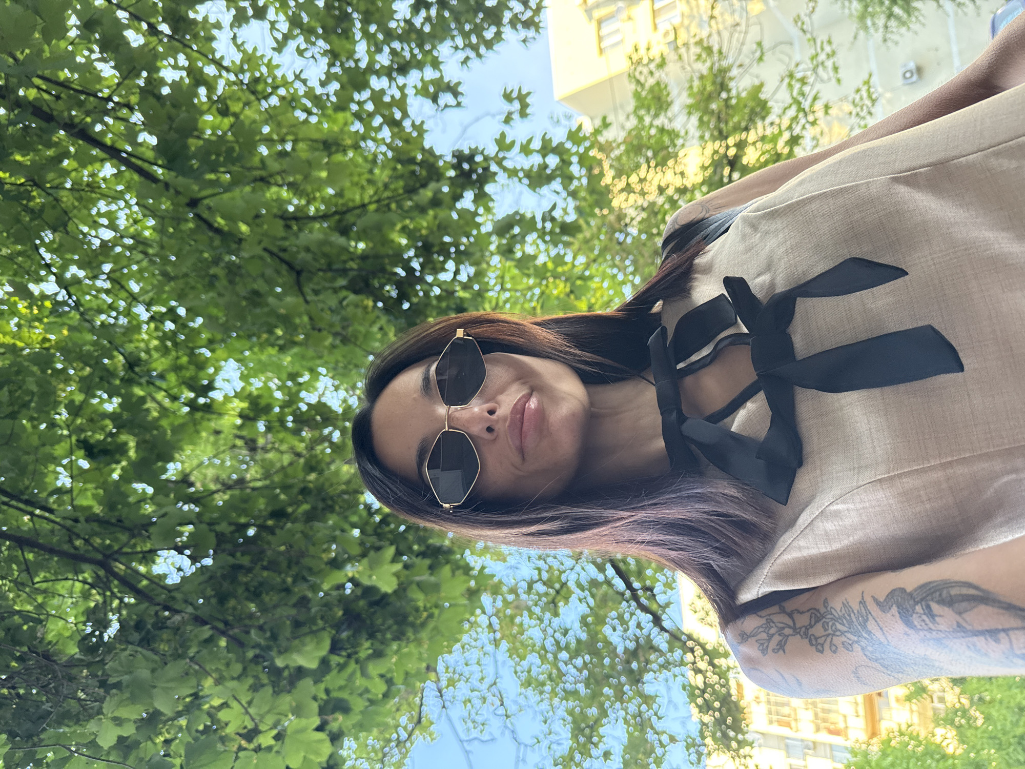 This is a low-angle portrait of a woman, captured outdoors in Pleven, Bulgaria. The image focuses on her head and upper torso, with her looking directly at the camera with a subtle smile or smirk. She has long, dark hair and is wearing distinctive hexagonal sunglasses with gold frames. Her attire consists of a light beige or off-white top adorned with multiple black ribbon bows on the front. Intricate tattoos, appearing floral or nature-inspired, are visible on her left arm and shoulder.
The background is dominated by a lush canopy of green trees, suggesting a park or tree-lined street setting. Through the leaves, patches of clear blue sky are visible, indicating bright, sunny daytime weather. A light-colored building, possibly yellow or cream, is partially visible in the upper right background, adding an urban element to the scene. The natural light brightly illuminates her features and the vibrant greenery. No text is visible in the image.