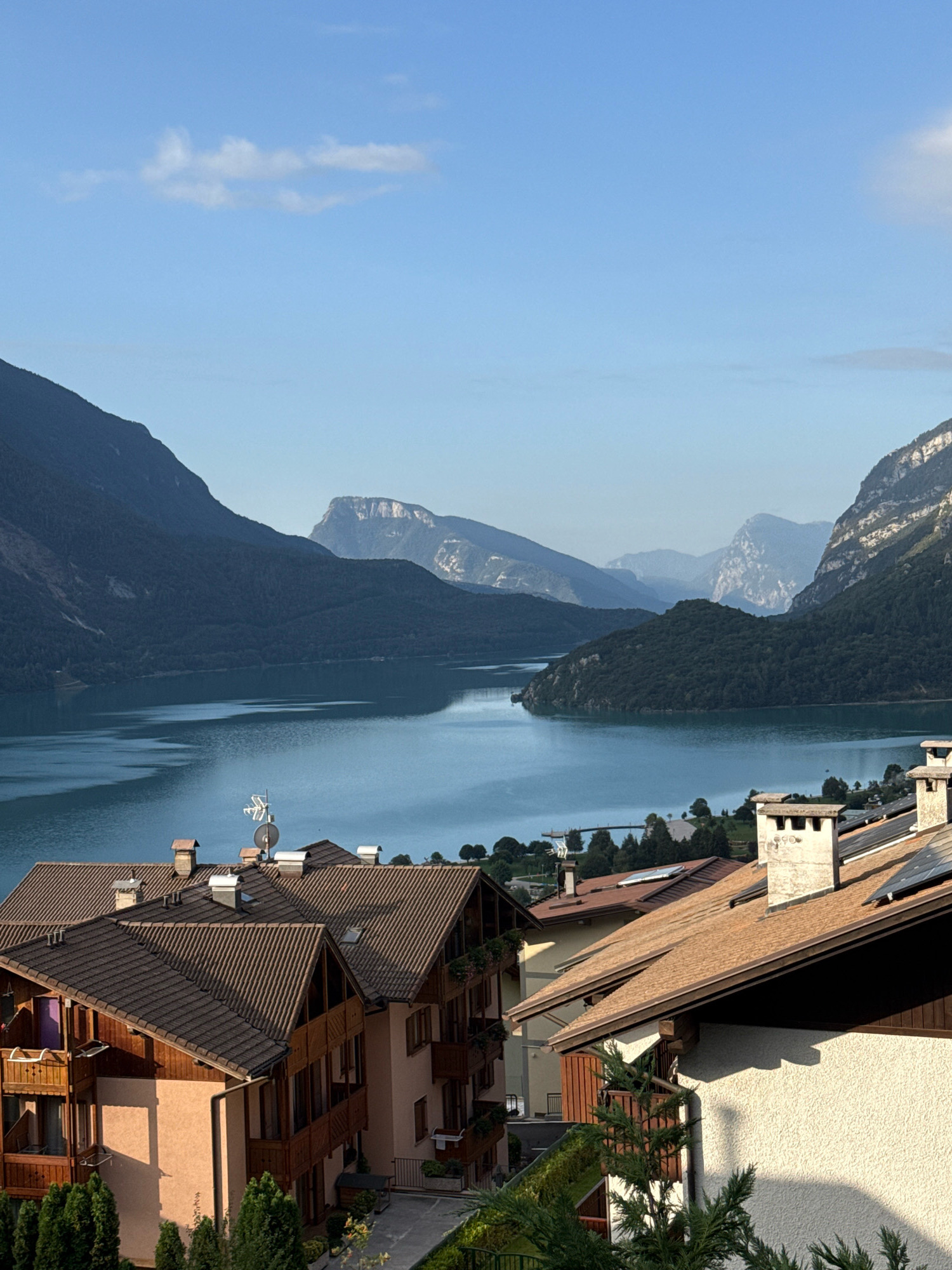 The image depicts a scenic view of the city of Molveno, Italy, with a focus on the picturesque lake and surrounding mountains. In the foreground, several residential buildings with characteristic tiled roofs and wooden balconies are visible. One building features a stucco facade, while another displays a peach-colored exterior. Chimneys and satellite dishes are present on the rooftops. Beyond the buildings, a vast, tranquil lake stretches out, reflecting the clear blue sky. The lake is bordered by lush green hills and imposing, dark blue mountains that dominate the background. The sky is bright blue with a few scattered white clouds. The overall scene suggests a peaceful, idyllic setting in a mountainous region.