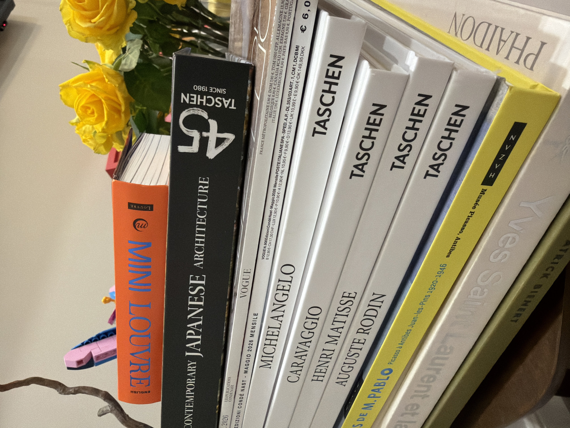 The image shows a stack of books of various sizes and colors, leaning slightly to the right. From left to right, the book spines read: "MINI LOUVRE," "CONTEMPORARY JAPANESE ARCHITECTURE TASCHEN SINCE 1980 45", "VOGUE Edizioni Condé Nast Maggio 2026 Mensile", "MICHELANGELO TASCHEN", "CARAVAGGIO TASCHEN", "HENRI MATISSE TASCHEN", "AUGUSTE RODIN TASCHEN", "Picasso à Antibes Juan-les-Pins 1920-1946 S DE M. PABLO", "Yves Saint", and "PHAIDON." There are yellow roses in the background and a wooden branch at the bottom left. A small blue and pink object is visible between the "MINI LOUVRE" and "CONTEMPORARY JAPANESE ARCHITECTURE" books. Text on the "VOGUE" book includes prices in various currencies. The books seem to be placed on a shelf.