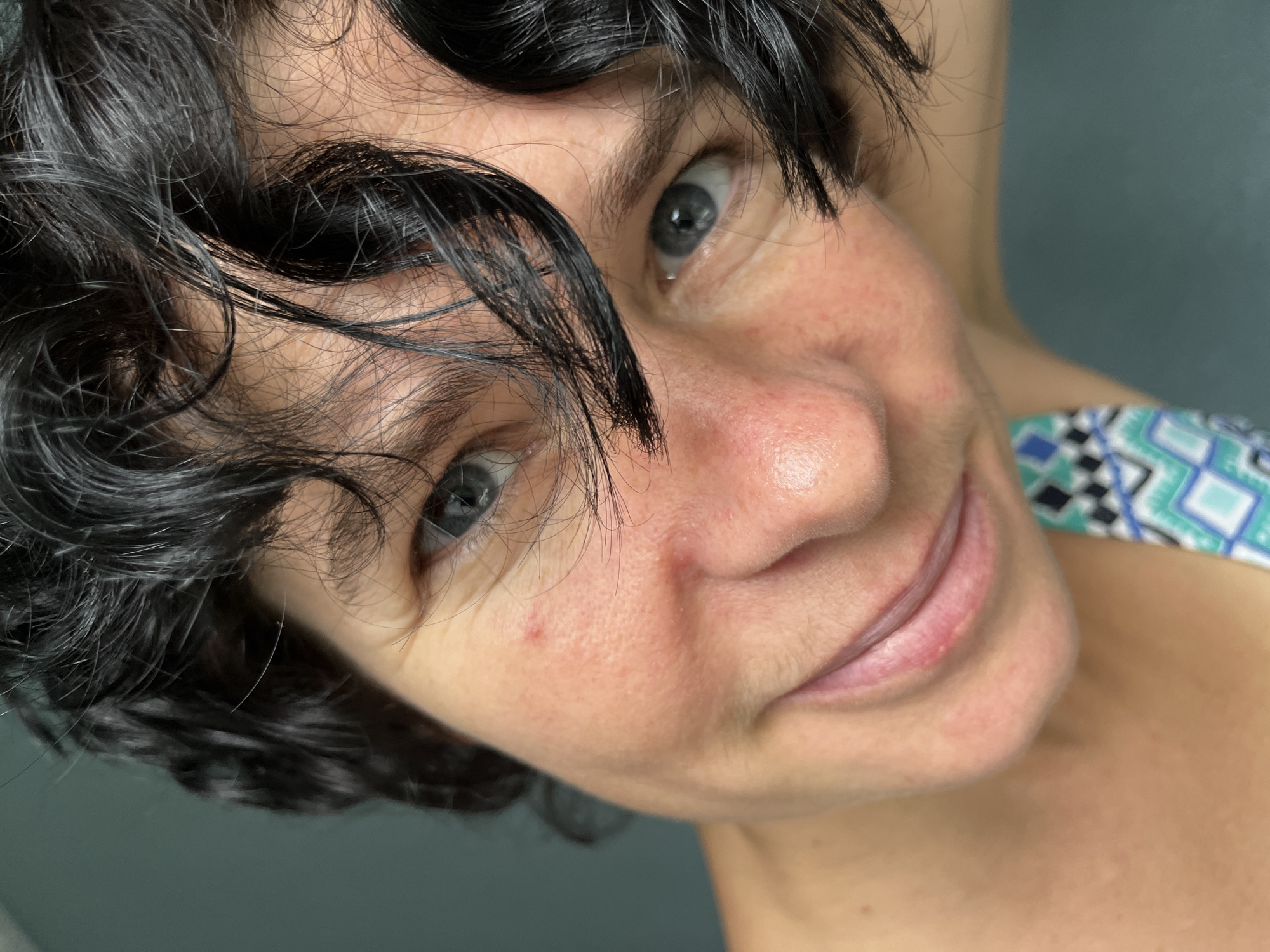 The image is a close-up, high-angle shot of a woman's face, cropped from the nose upwards, with her head tilted back. She has dark, curly hair that falls across her forehead and partially obscures her face. Her skin is fair with some redness and visible pores. She has grey-blue eyes and a slight smile. A faint mole is visible on her cheek. Her neck and part of her shoulder are visible, and she is wearing a top with a blue, white, and green geometric pattern. The background is a plain, dark teal wall. There is no discernible context to indicate a specific location in the city of ღრმაღელის თემი, Georgia.