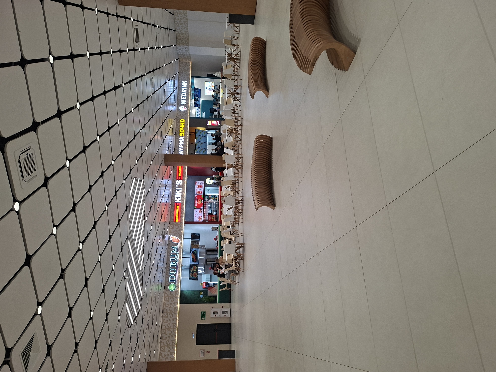 This is an interior shot of a food court in Kokshetau, Kazakhstan, likely in a shopping mall. The scene showcases several fast-food establishments, including "DURUM," "KIKI'S," "AYPMA SOЧHO," and "WEDRINK." The perspective is from an elevated viewpoint, looking down the length of the food court.
The environment is characterized by a geometric ceiling pattern with rounded squares connected by black lines and small lights. The floor appears to be tiled in a neutral tone. There are several unique, curvilinear wooden benches for seating. Tables and chairs are arranged in front of the food stalls.
People are seen dining at the tables. The food court features brightly lit signage for the various vendors.