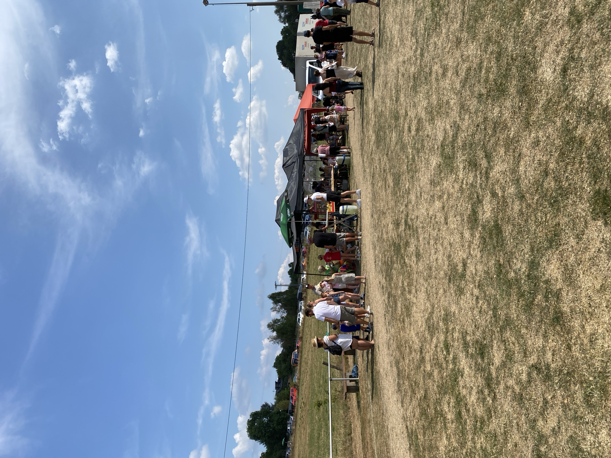 A wide-angle shot captures a daytime outdoor gathering at a grassy field, likely a community event, under a clear blue sky with scattered white clouds. In the foreground, dry, yellowish grass covers the ground. In the middle ground, a group of people are gathered around several vendor tents. The tents are black and orange, with one green tent to the left. Some people are standing, talking, and looking at items on display. A few children are also present, with one young girl running in the middle of the frame. Behind the tents, a white truck with the text "VOLKSWAGEN" is visible, along with other vehicles parked in the background on a hill. The location is identified as Ingelbach, Germany. The overall atmosphere suggests a relaxed, possibly festive, event.