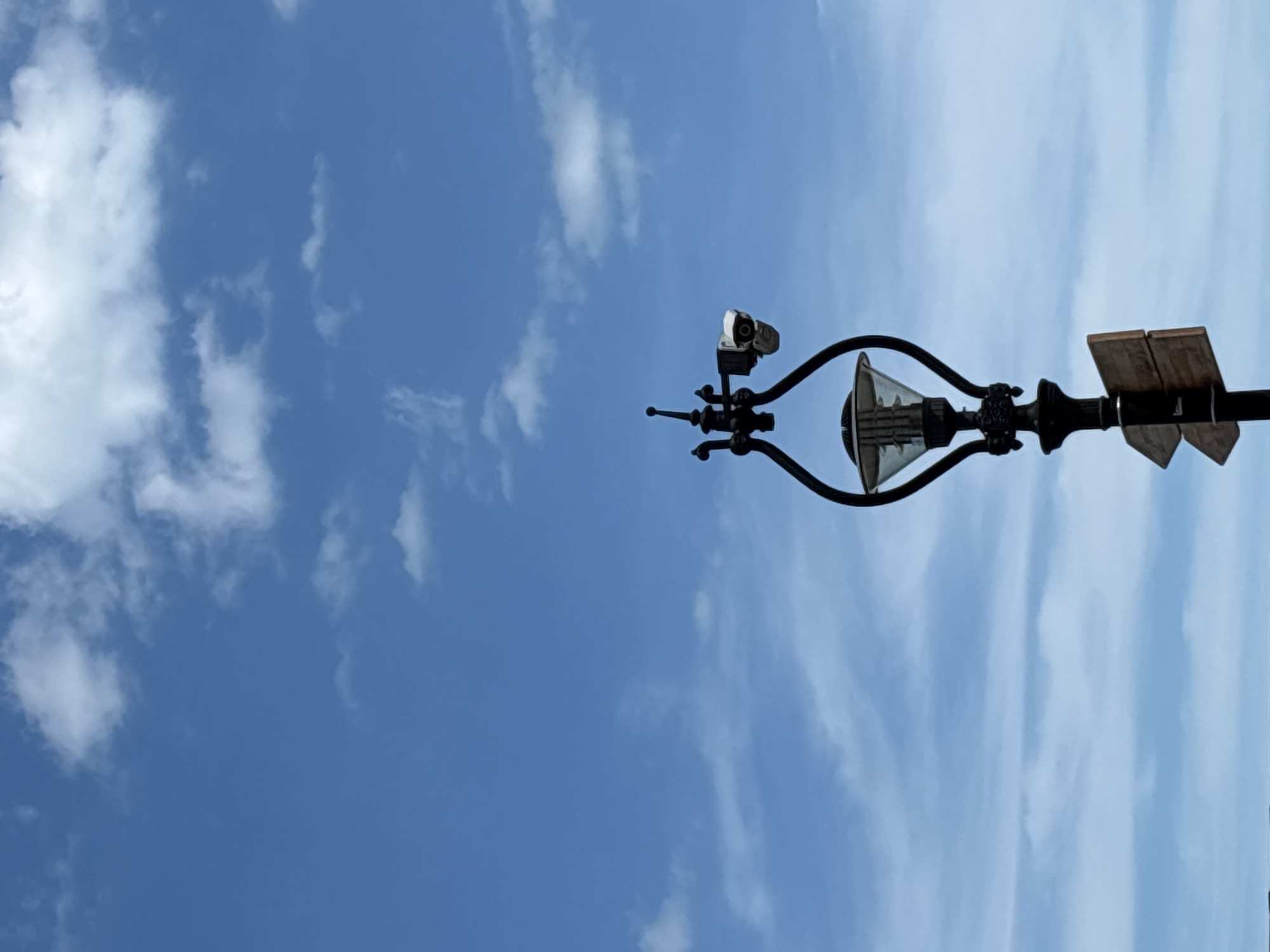The media file shows a streetlamp against a bright blue sky with scattered white clouds. The streetlamp is a dark, ornate metal fixture with a glass lantern and a camera attached to its top. There are also two wooden panels on top of the streetlamp. The image is captured from a low angle, looking up at the sky. The lighting suggests it is daytime, possibly late morning or early afternoon. The weather appears clear and sunny with a few wispy clouds. No people are visible in the frame, and the focus is entirely on the streetlamp and the sky. The presence of the camera on the streetlamp might indicate a public area or a place where surveillance is in effect. The overall scene conveys a sense of an ordinary day outdoors in an urban or suburban environment, with the streetlamp being a prominent man-made object against the natural backdrop of the sky.