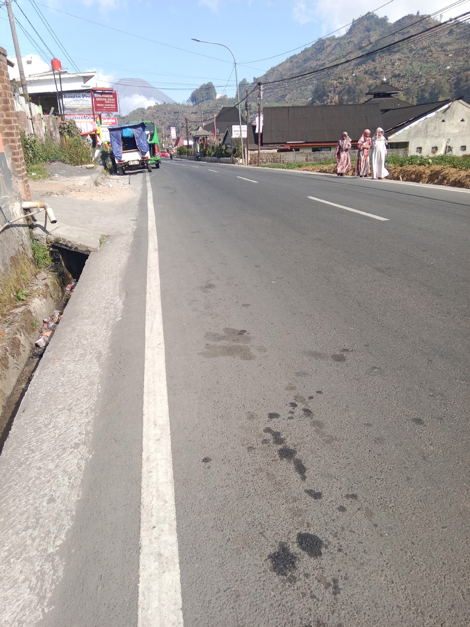 This image captures a street scene in Parikesit, Indonesia, under a clear blue sky. The road, paved with asphalt and marked with a white line, extends into the distance. On the left side of the road, a concrete drain is visible, containing debris and several discarded cans. Adjacent to the drain is a dirt embankment with some vegetation. A few buildings line the right side of the road, one with a dark roof and another painted white. In the foreground, there are dark oil stains on the asphalt. In the middle ground, three women wearing traditional clothing walk along the side of the road. Further down the road, a green and white motorized vehicle is parked on the left, and a few other vehicles are visible. In the background, a mountain rises, with terraced slopes and a plume of smoke or mist emanating from its peak. Power lines crisscross the sky. Signs advertising "Warung Pak Kumis" are posted on buildings on the left side of the street.
