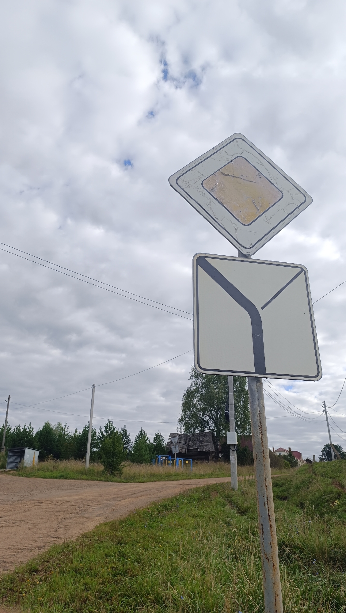 This is a medium shot, outdoors, low-angle vertical photograph of a street sign at a crossroads in Karsha, Russia. The sky is overcast and cloudy, with patches of blue visible. The street sign consists of two signs mounted on a pole. The upper sign is a diamond shape with a white border and a yellow interior, a "Main Road" sign. The lower sign is a square with a white border and a dark grey line indicating a yield or turn. The road is unpaved, with dirt and grass. In the background, there are trees, a dilapidated wooden building, and some houses. There are no people or animals visible in the photograph. The overall scene depicts a rural intersection.