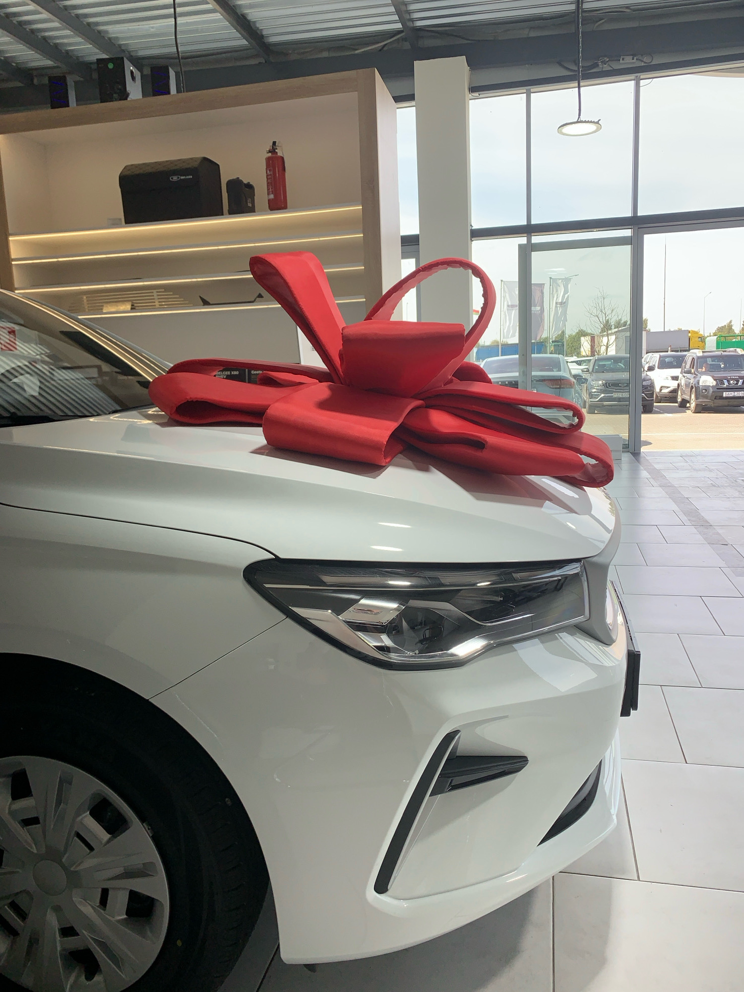 A white car is parked inside a showroom, adorned with a large red bow on its hood, suggesting a gift or promotional display. The car's front is visible, showing the headlight and bumper. In the background, through large glass windows, other cars are parked outdoors, some with visible license plates. The showroom floor is tiled, and the ceiling has industrial lighting. Shelving units with various items, including what appears to be a black case and a fire extinguisher, are mounted on the wall. The overall scene depicts a car dealership environment.