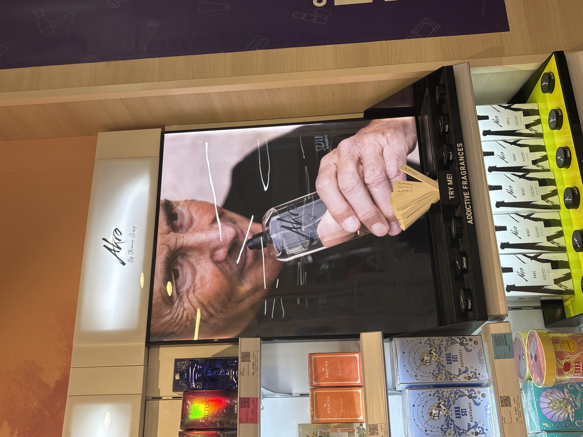 This appears to be a photograph taken inside a retail store, likely a department store or perfumery, in Perm, Russia. The image showcases a display for the Akro brand of fragrances by Olivier Cresp, alongside other fragrance brands such as Anna Sui.
The central focus is a display screen showing a close-up image of a person holding an Akro fragrance bottle, presumably smelling it, with white lines added to the image. To the right of the screen is a shelf with Akro fragrance samples labeled with words such as "Smile" and "Night."
In the lower portion of the image, there are shelves displaying products from other brands, including Ajmal Evoke and Anna Sui Fantasia fragrances. Price tags in Russian currency (P) are visible. The environment seems well-lit and organized. The presence of different fragrance brands indicates a competitive retail setting.